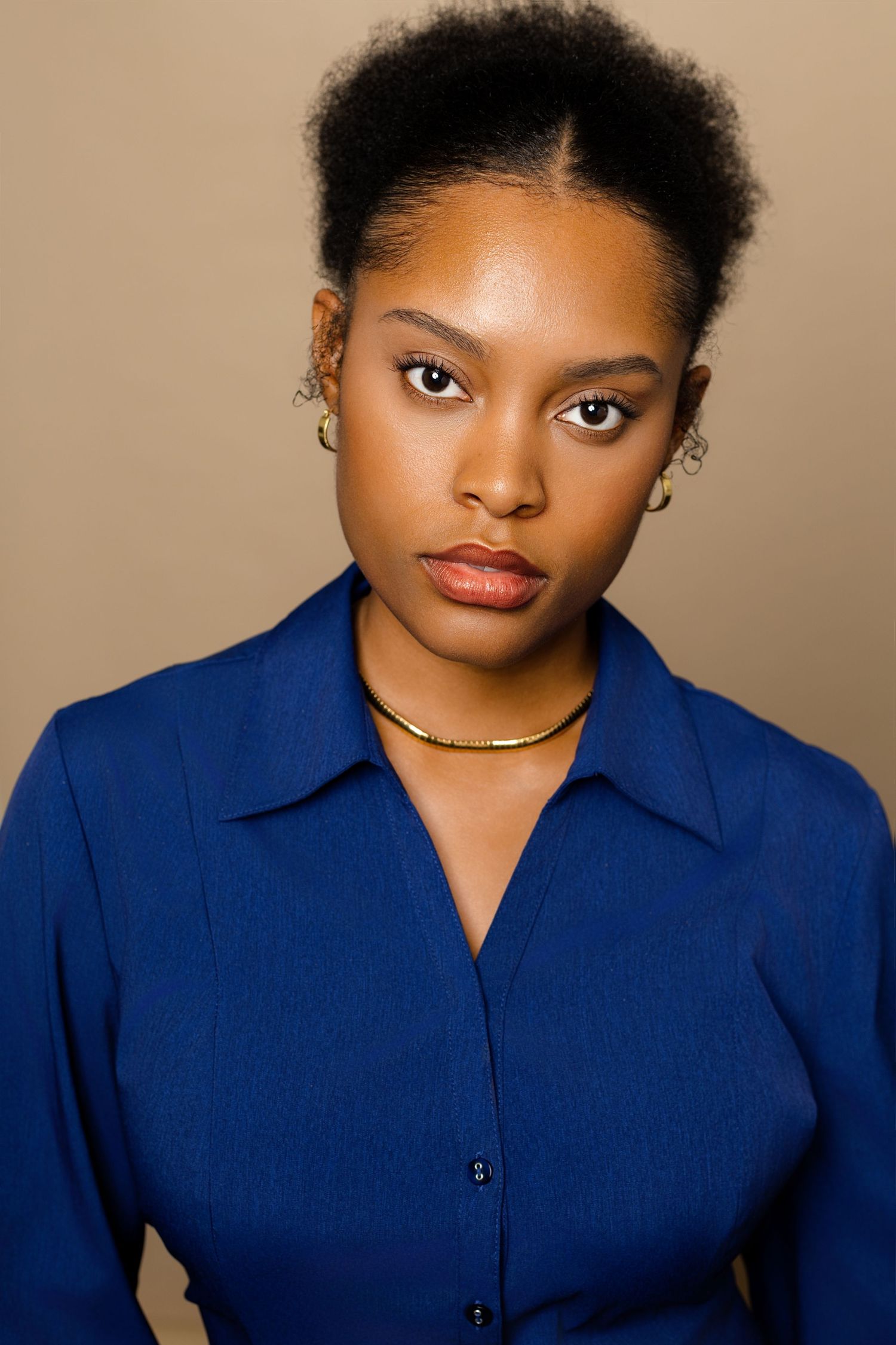 Young black woman in blue shirt with gold jewelry poses for professional headshot on tan background.