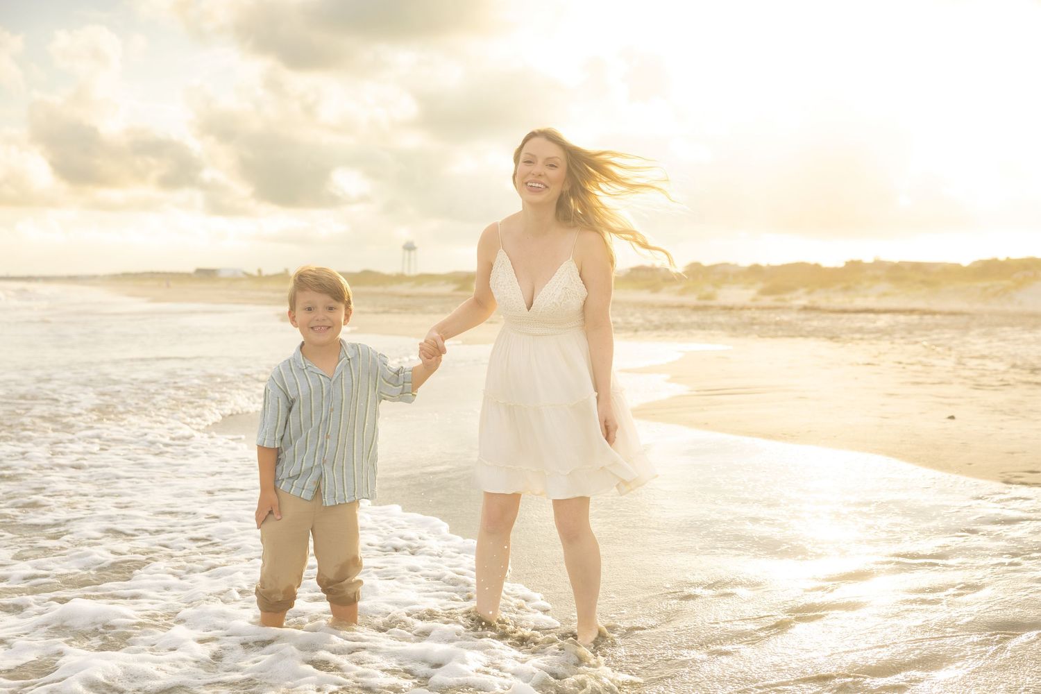 A woman in a white dress holds hands with a young boy on a sunny beach with golden light and ocean waves.
