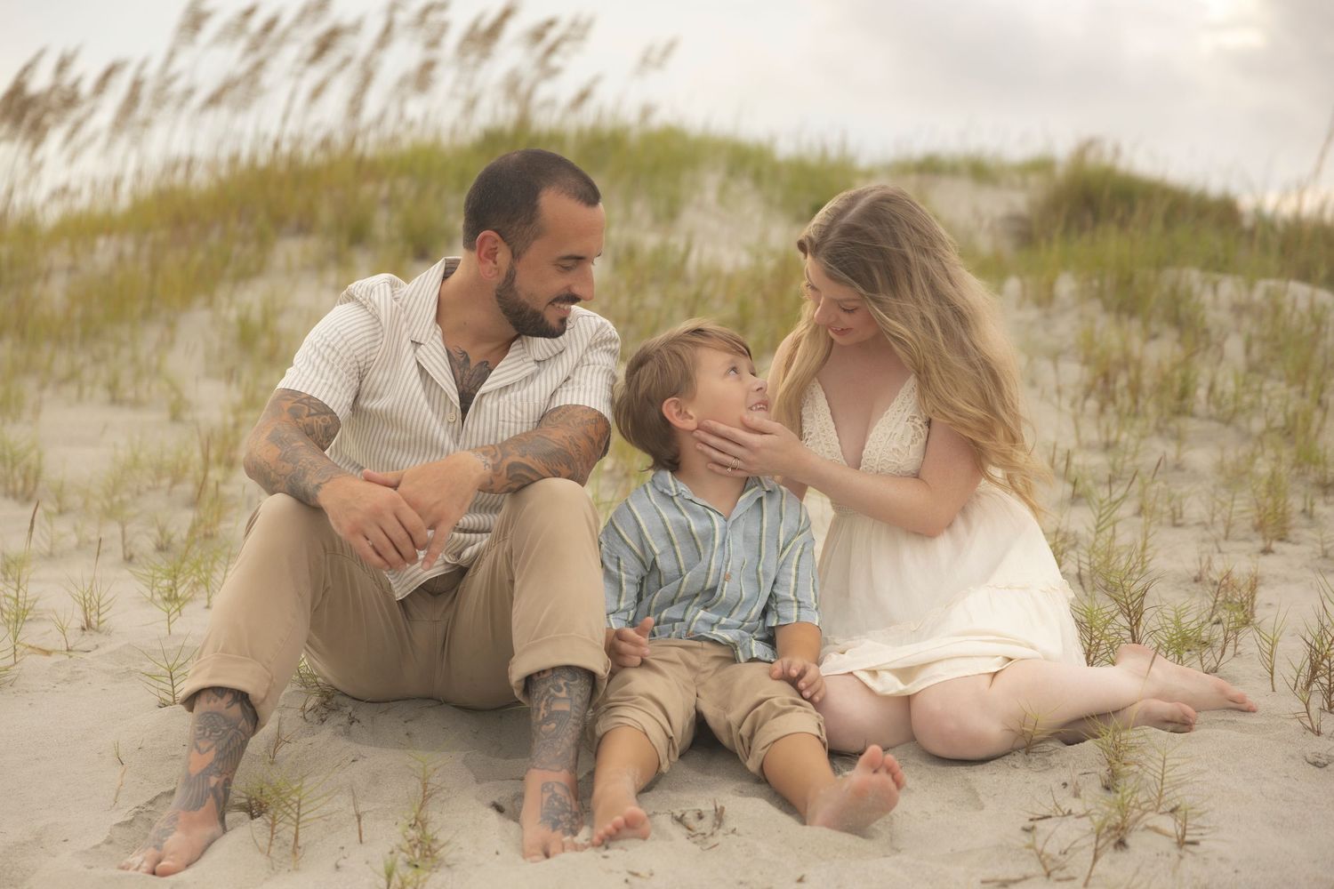 Family of three sitting on sandy beach dunes, parents smiling at young boy, casual summer attire, coastal setting.