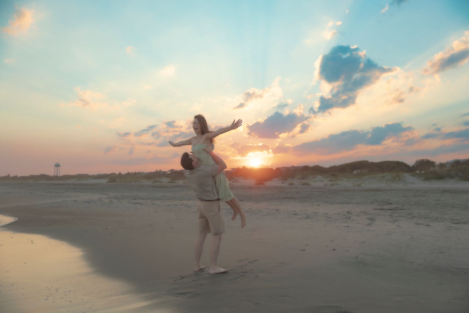 Joyful couple on beach at sunset, man lifting woman with arms outstretched against colorful sky.