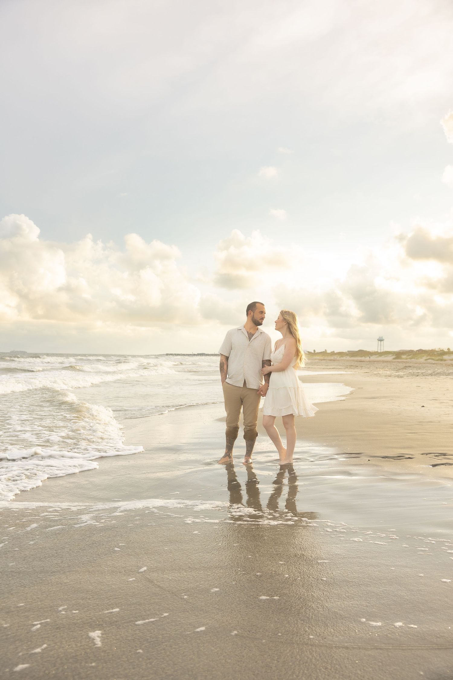Couple holding hands and gazing at each other while standing in ocean waves at sunset.