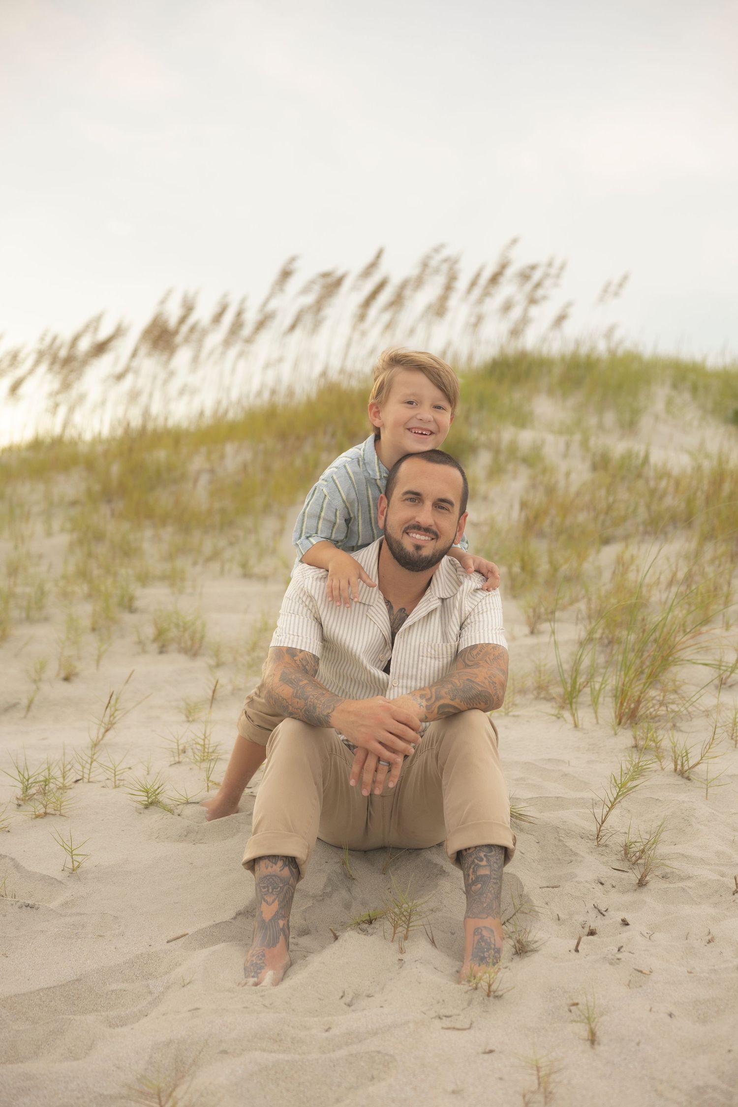 Tattooed father sitting on sandy beach dunes with young son hugging him from behind, both smiling.