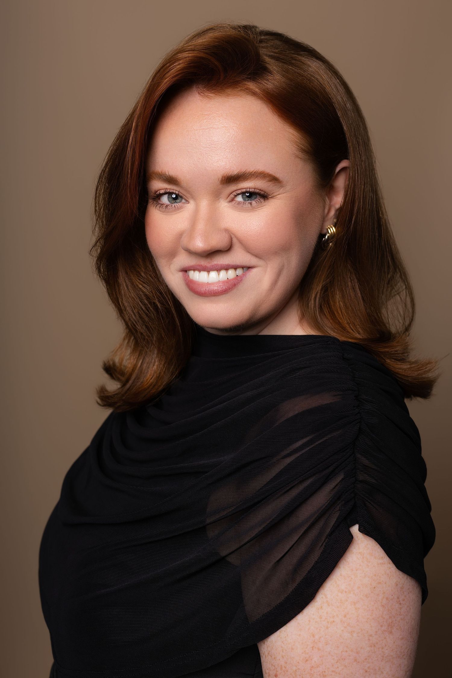 Smiling woman with auburn hair wearing a black off-shoulder dress against a taupe background.