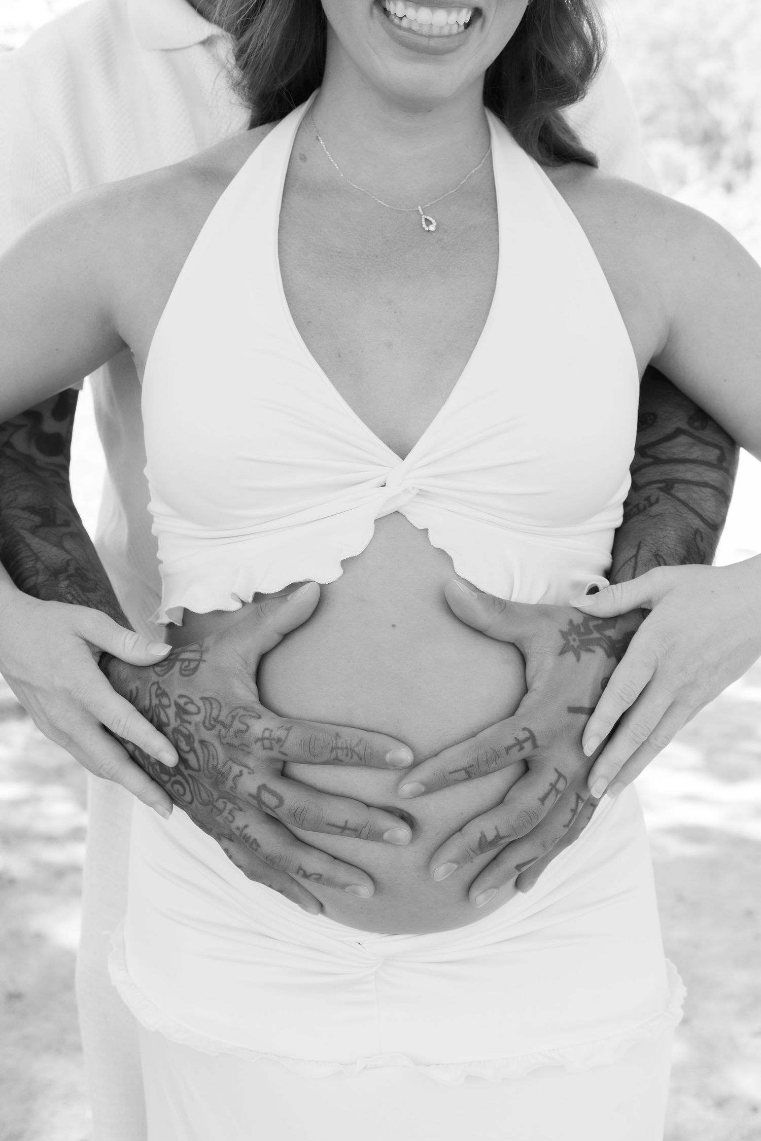 Black and white photo of a pregnant woman in white top with tattooed hands cradling her baby bump.