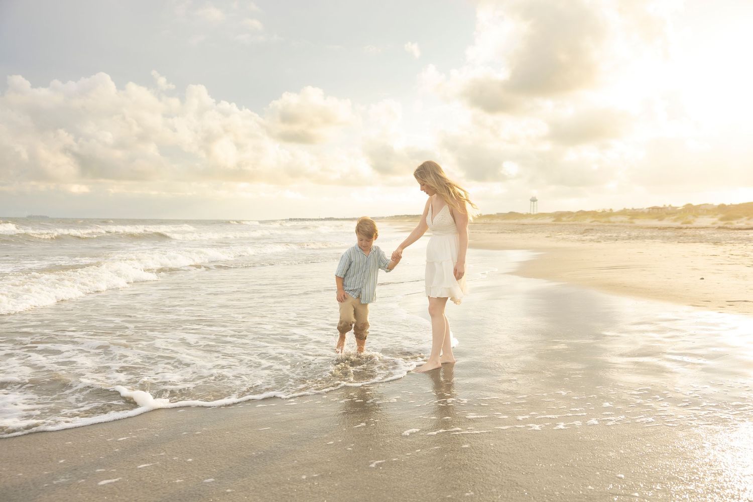 A mother and young child hold hands while walking along a sunny beach shoreline with gentle waves.