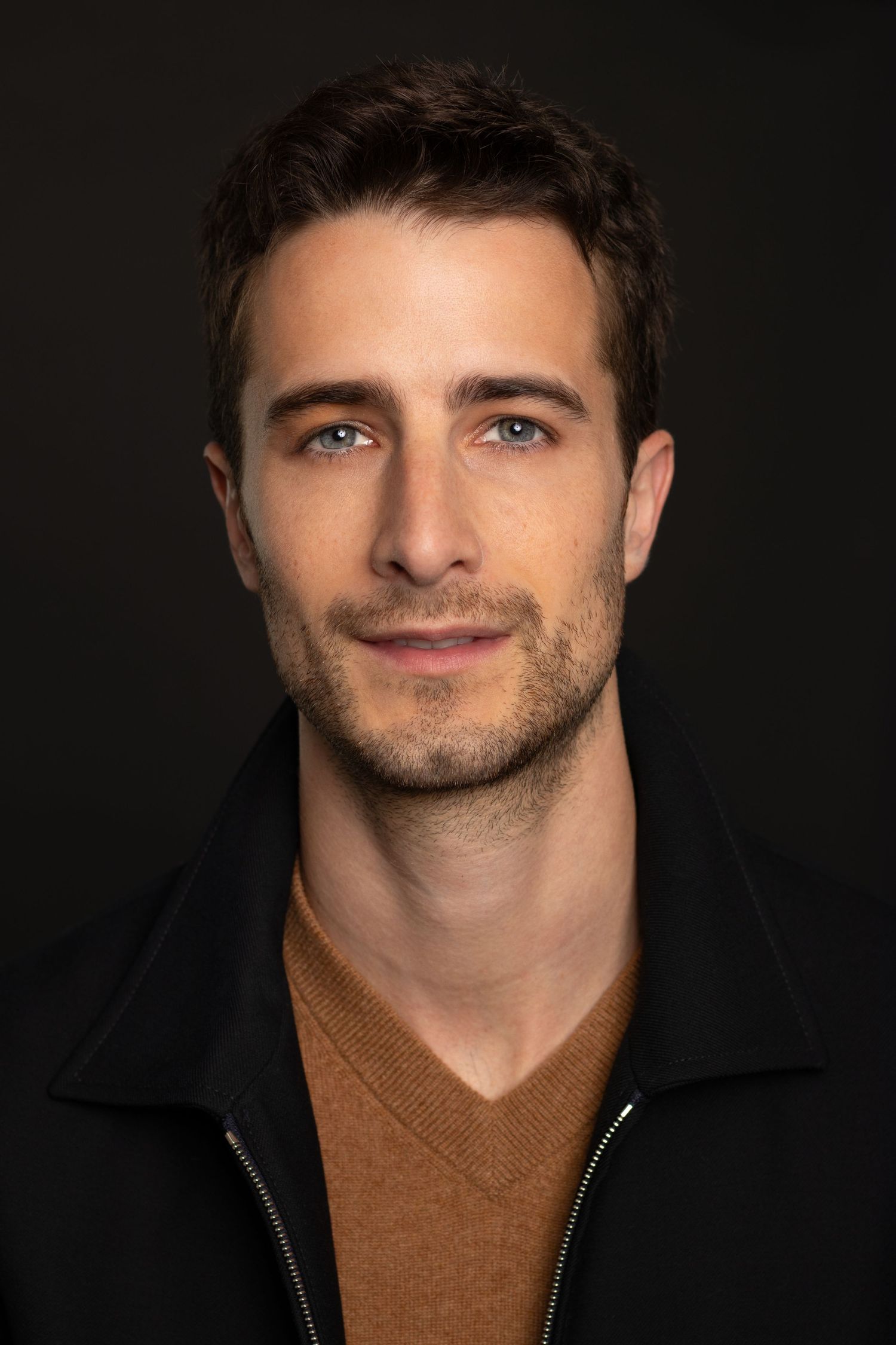 Handsome young man with blue eyes and stubble wearing a brown sweater and black jacket against dark background.