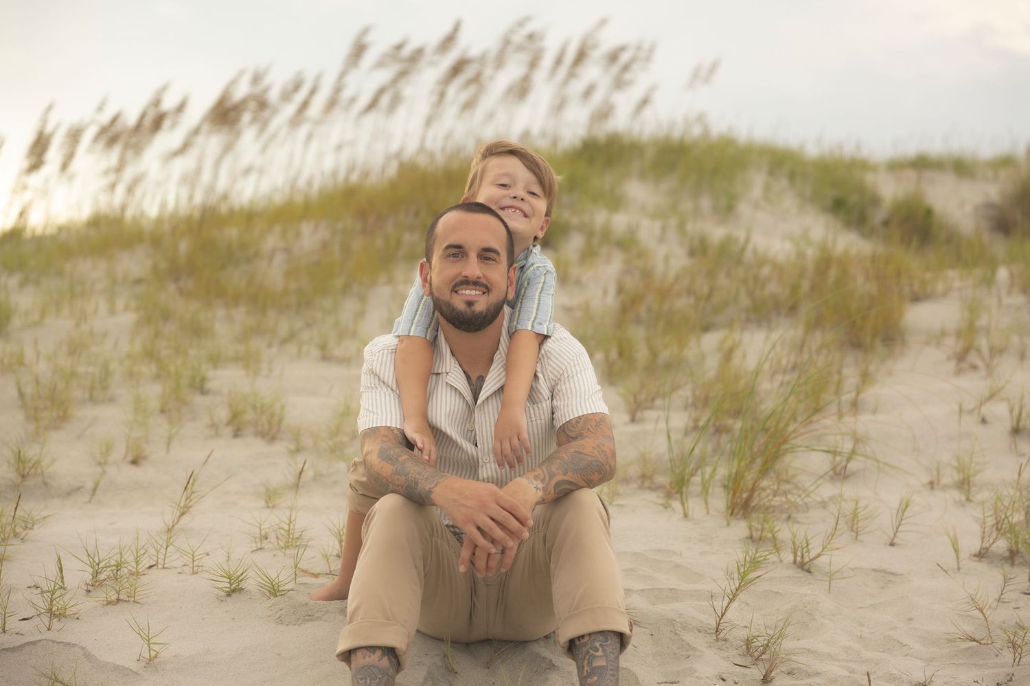 A man and young boy sit together on sandy beach dunes with sea oats in the background.