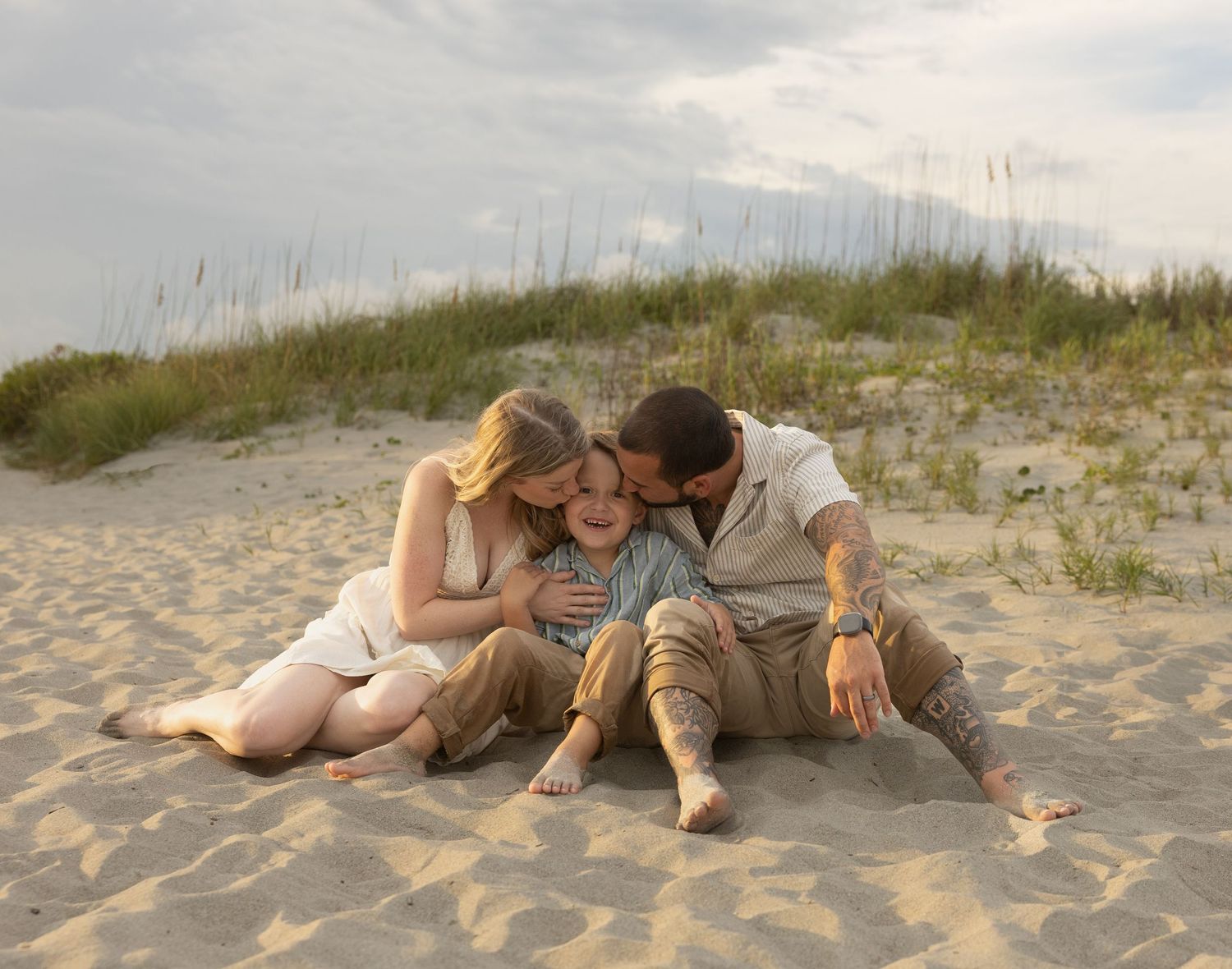 Family of three sitting on sandy beach dunes, parents kissing child on cheeks, casual outdoor portrait.