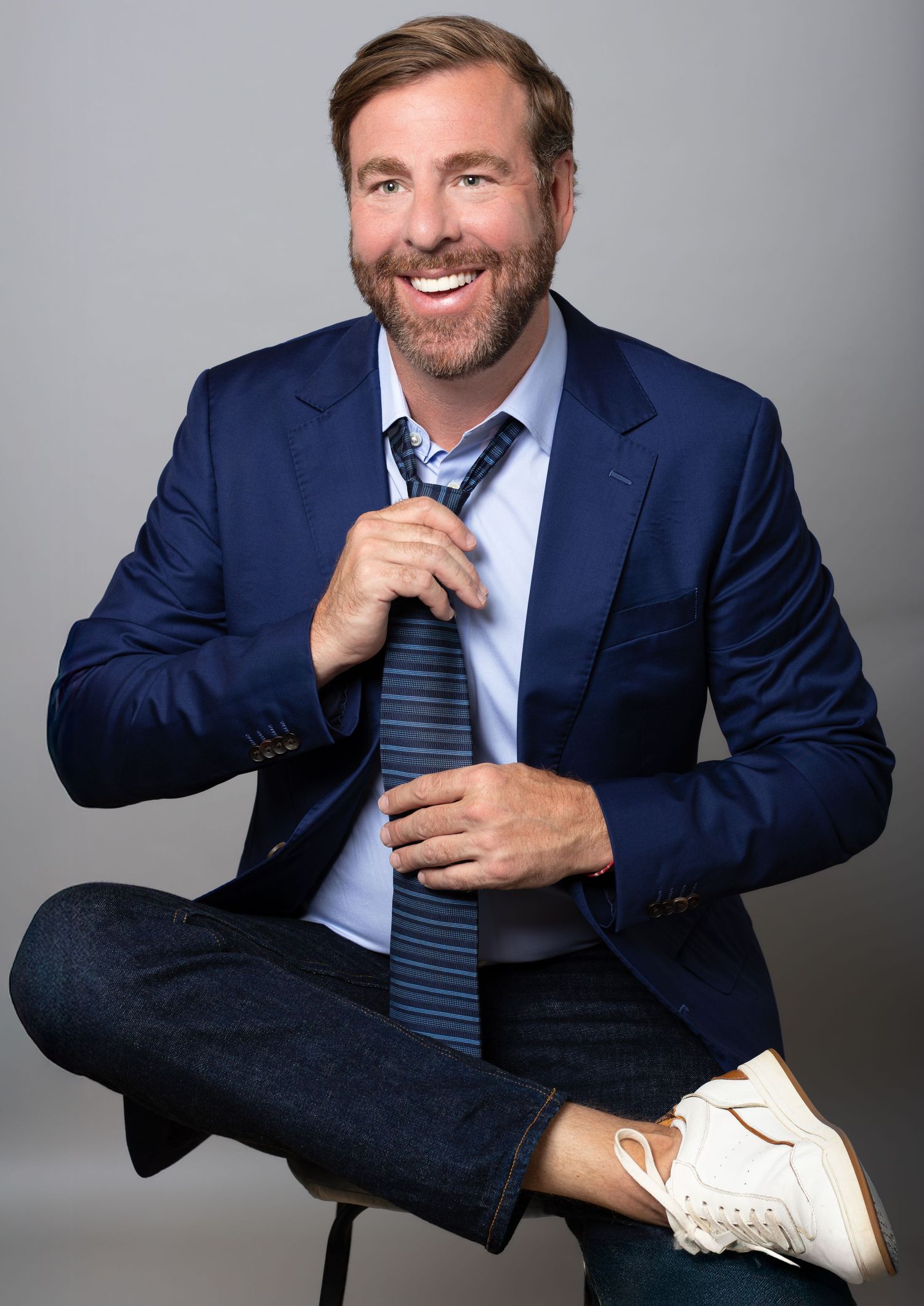 Smiling man in navy blazer and tie sits cross-legged on stool, wearing jeans and white sneakers, against gray background.