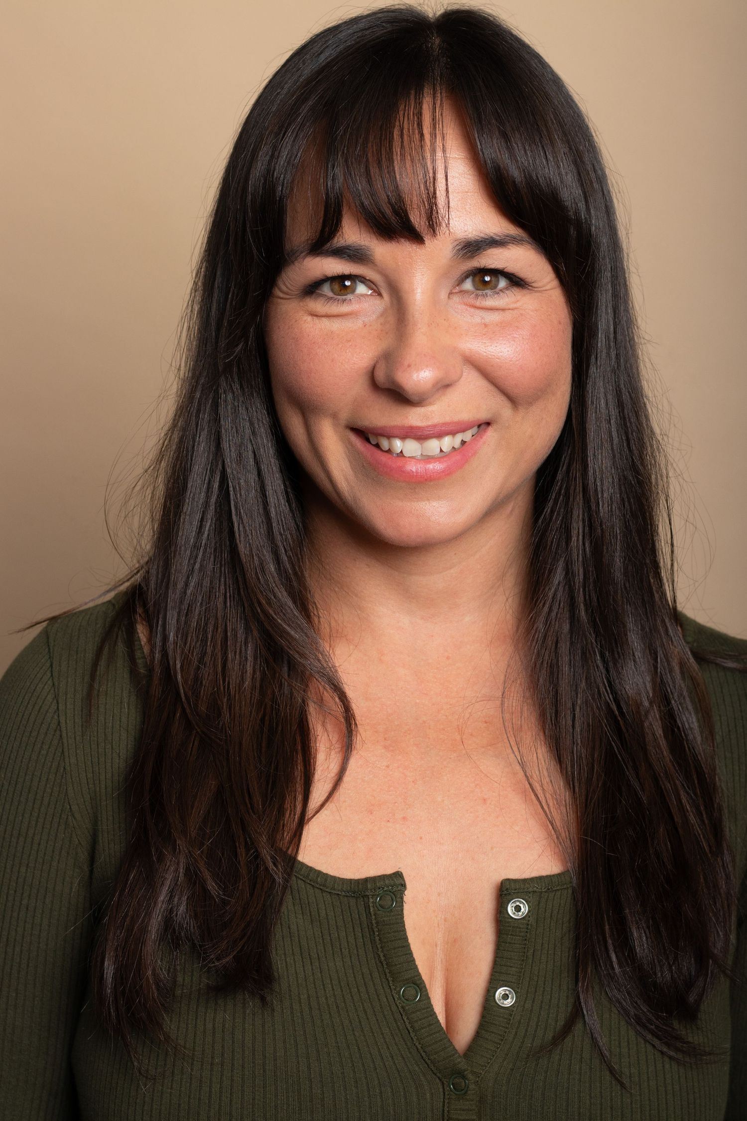 Smiling brunette woman with bangs wearing green top posing for portrait against beige background.