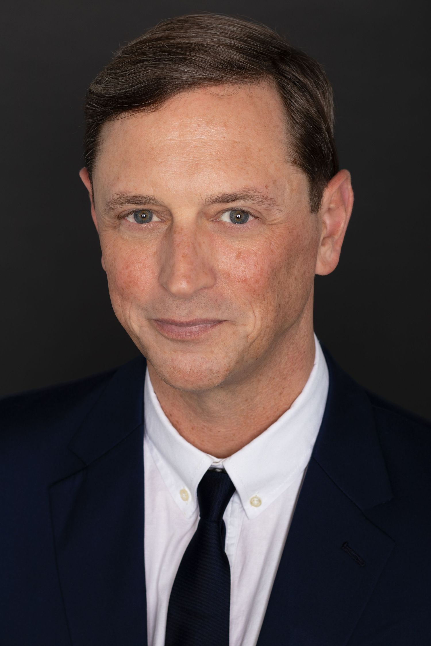 Middle-aged man in navy suit and tie poses for professional headshot against dark background.