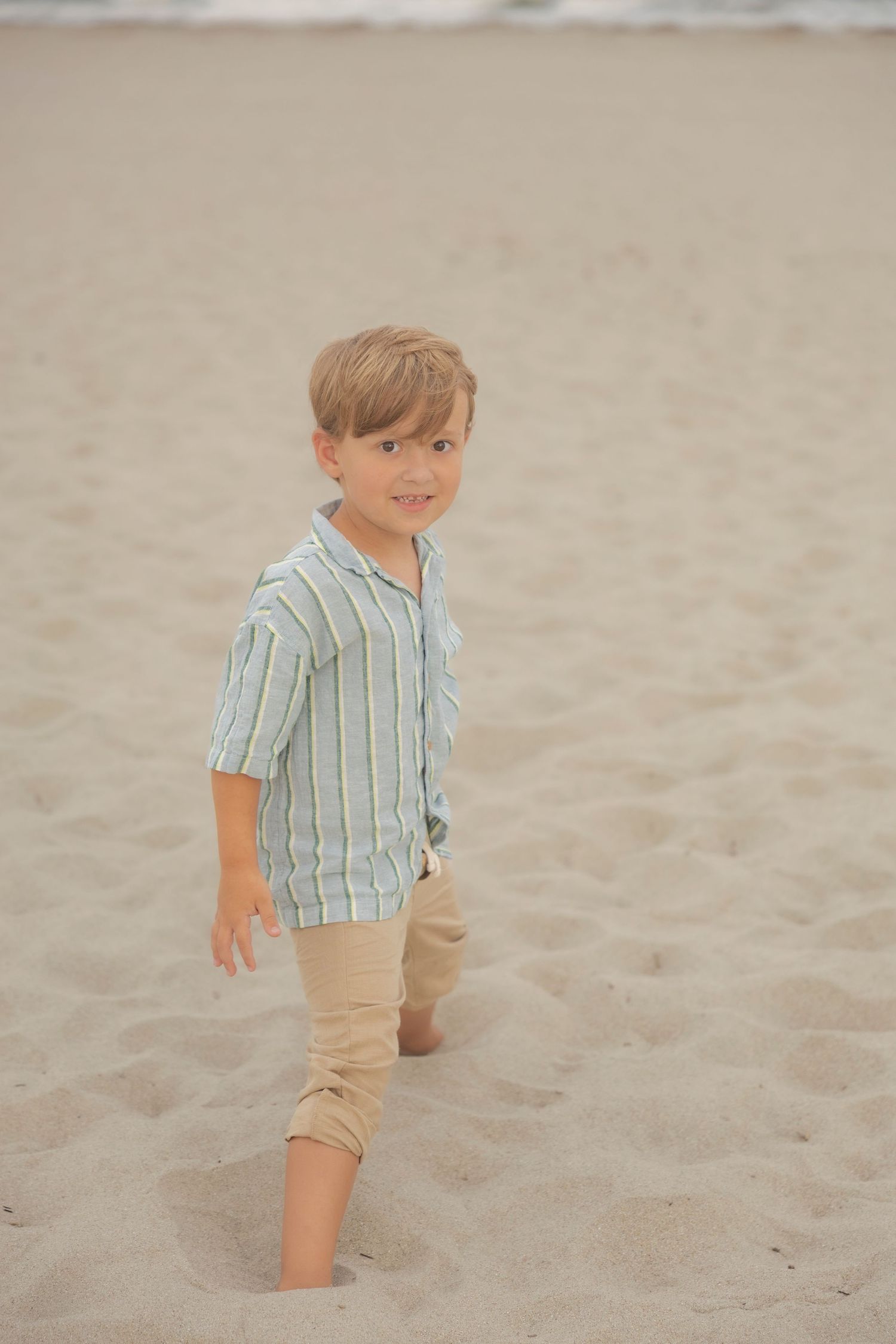 A young boy in a striped shirt and khaki pants walks barefoot on a sandy beach, smiling at the camera.