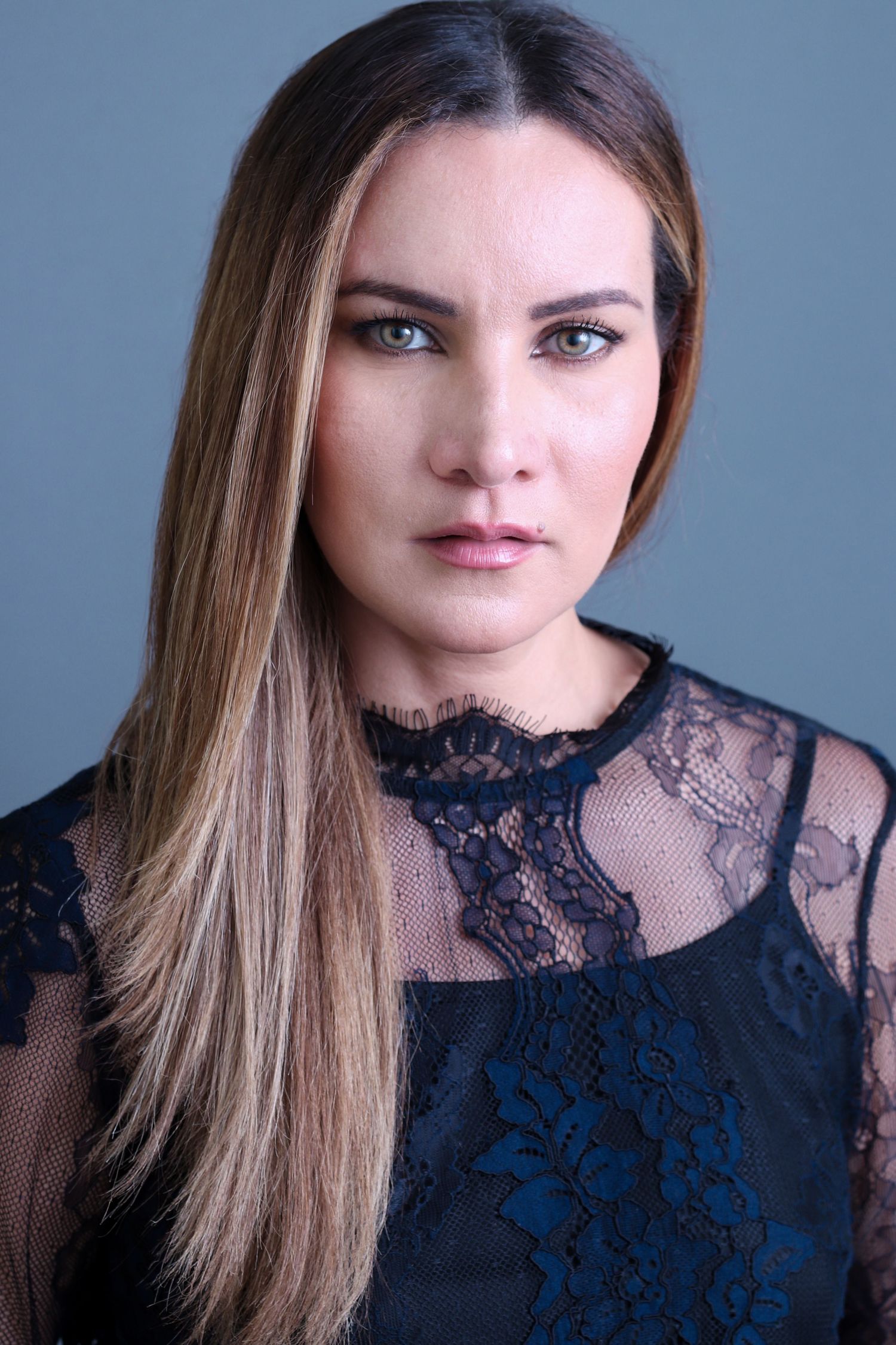 Woman with long brown hair in black lace top poses for professional actor headshot on blue background in Charleston, SC.