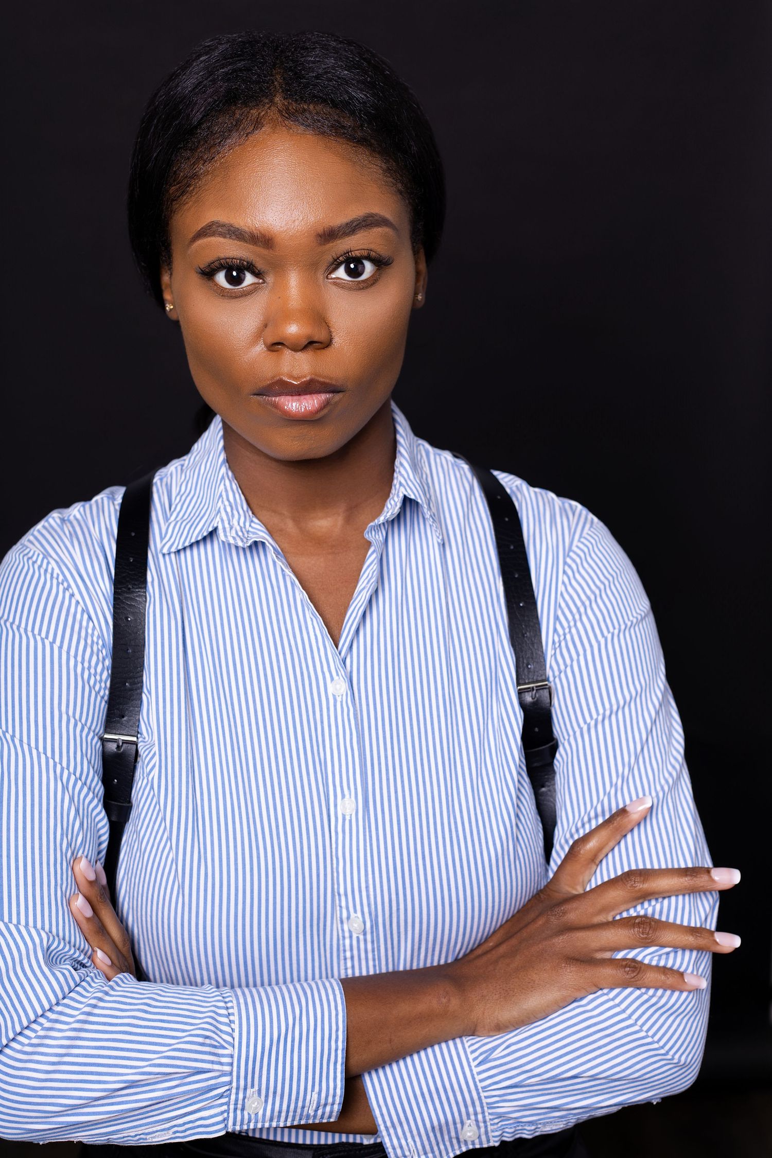 Black woman in striped blue shirt with suspenders poses with arms crossed for professional headshot in Charleston, SC.