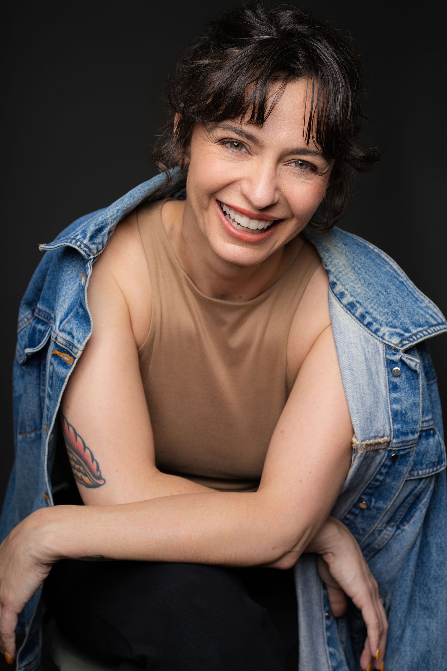 Smiling brunette woman in denim jacket and beige top with arm tattoo poses against dark background.