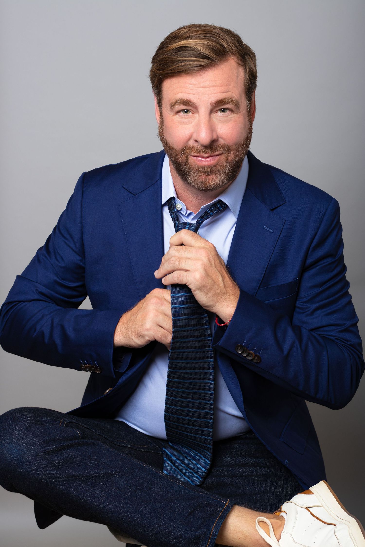 Smiling bearded man in a blue suit adjusts his tie while sitting cross-legged for a professional studio portrait in Charleston, SC.