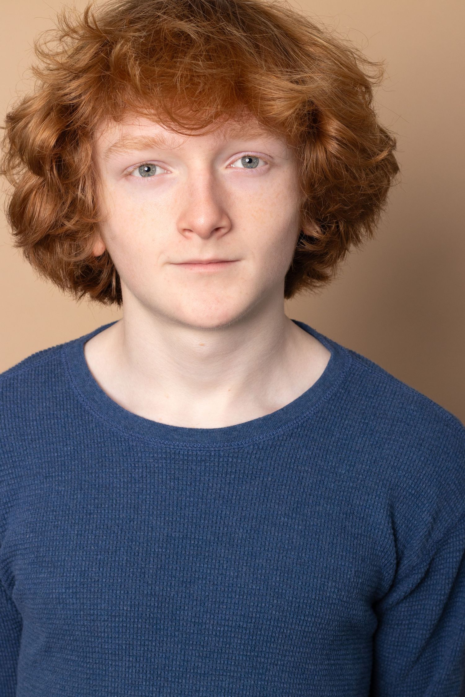 Young redhead teen with curly hair wearing a blue textured sweater against beige background.
