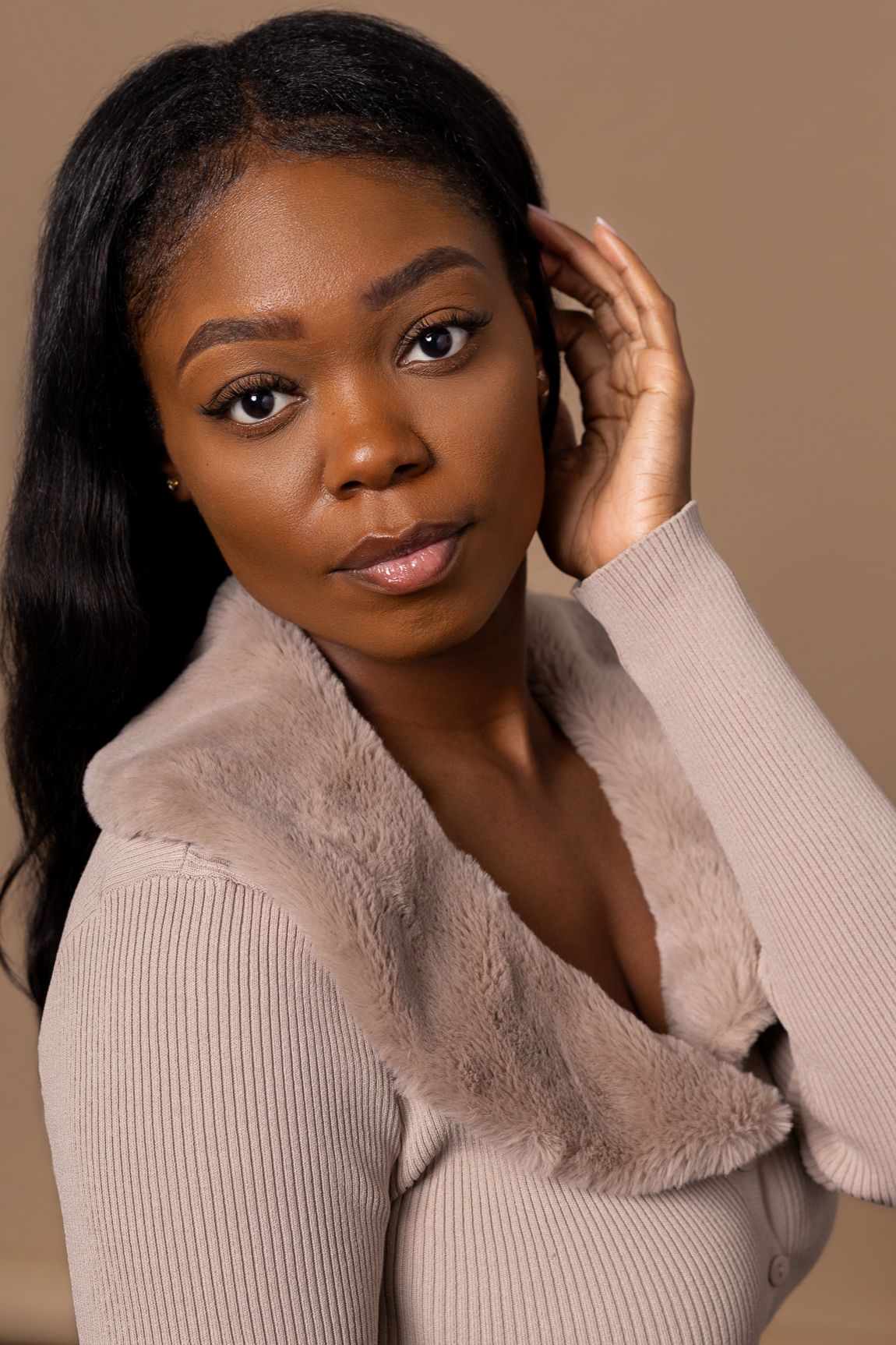 Black woman in beige fur-collar sweater poses elegantly for professional portrait on tan background.