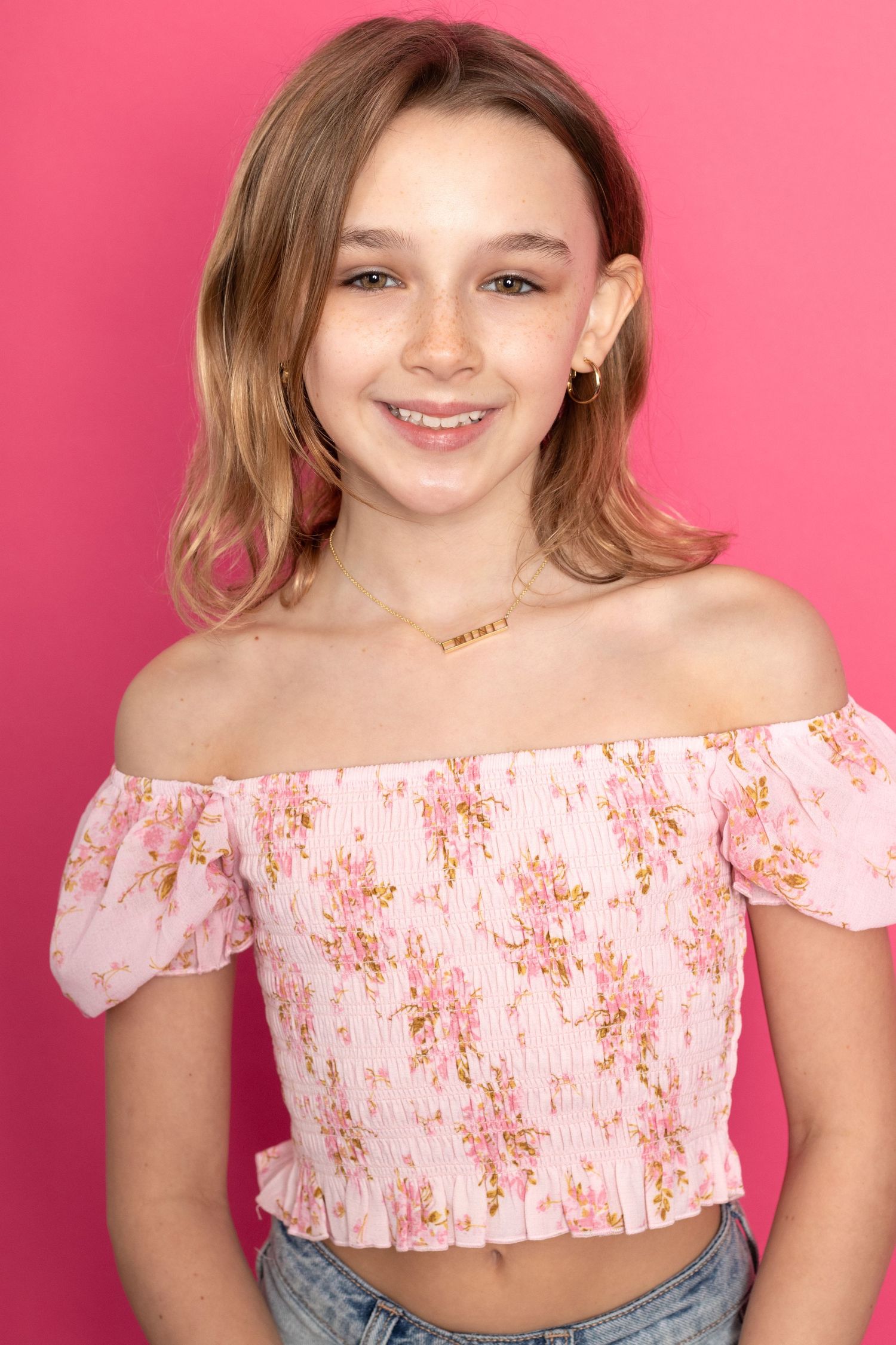 Smiling young girl with blonde hair wearing a pink floral off-shoulder top poses against a pink background.