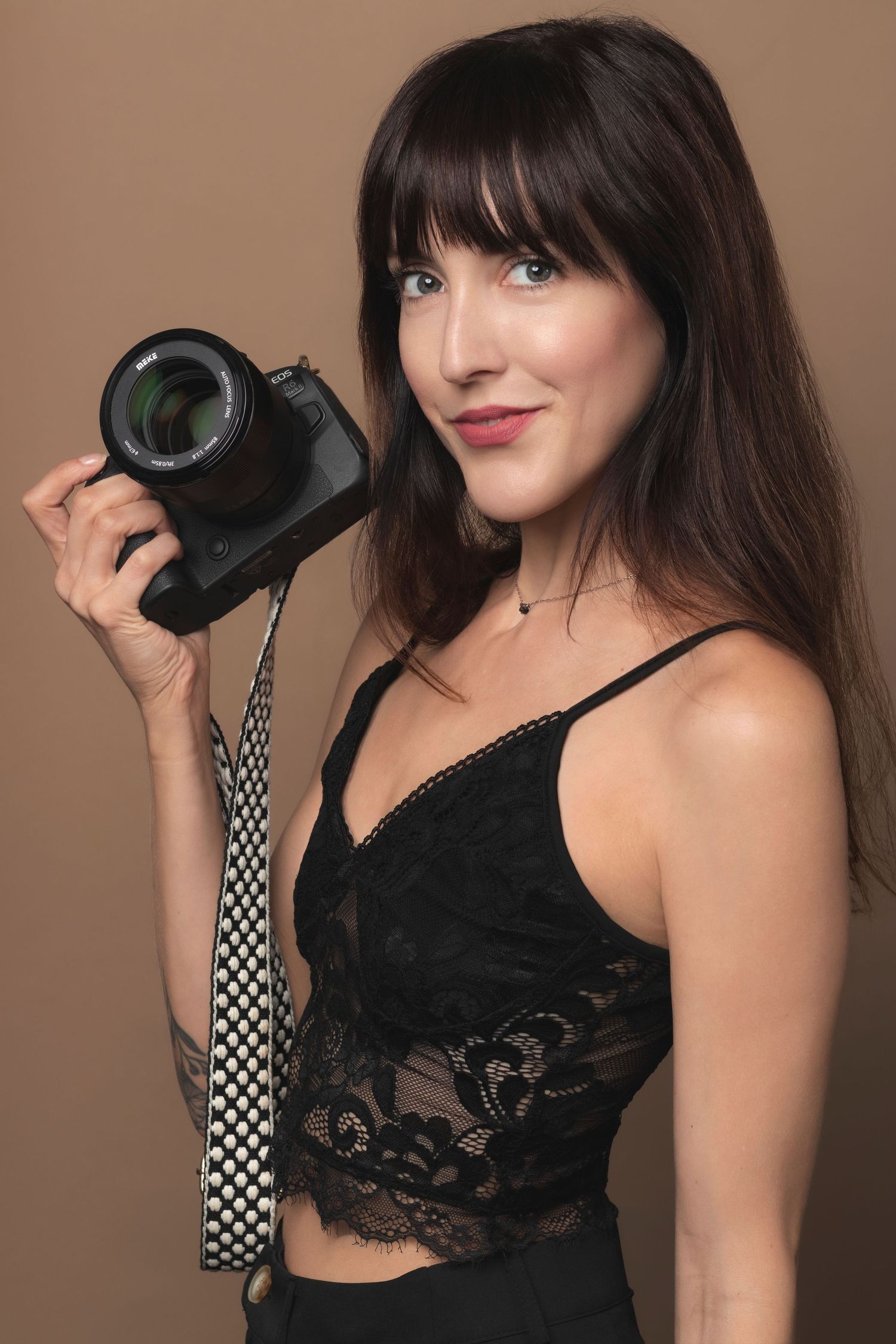 Brunette female photographer smiling while holding a DSLR camera against a tan background.