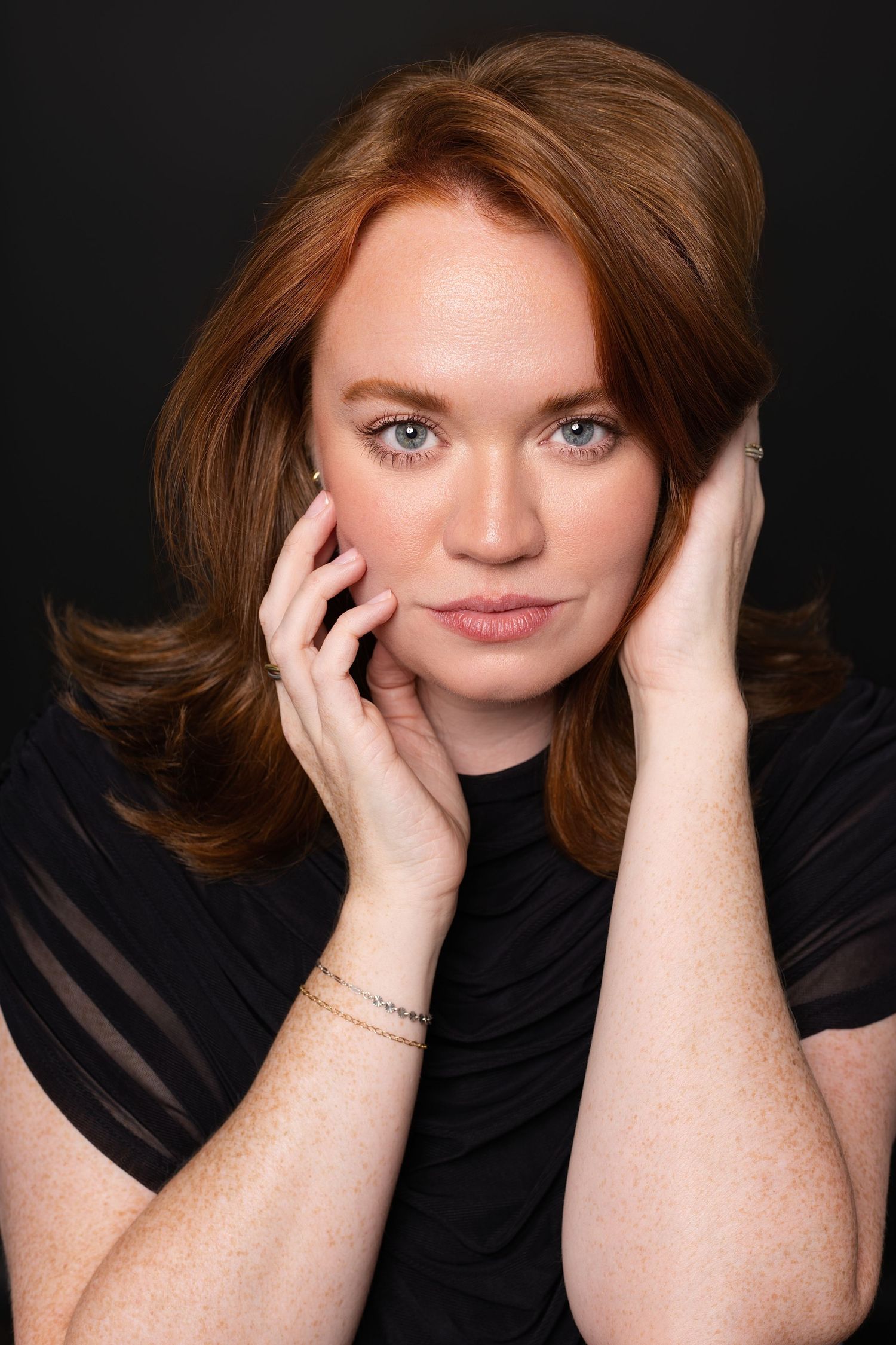 Red-haired woman with freckles and blue eyes poses with hands framing face, wearing black top on dark background.