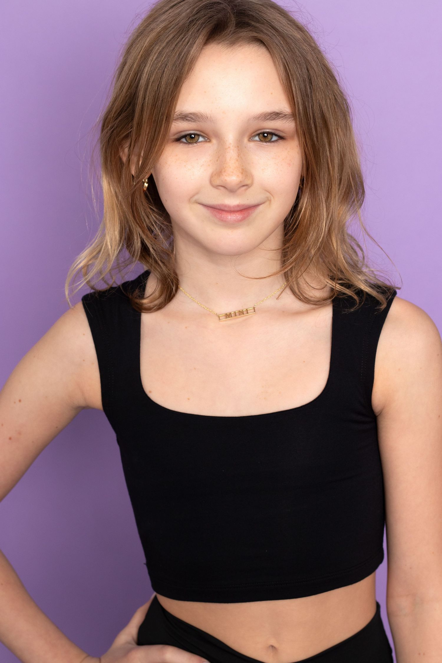 Young woman in black crop top and necklace posing against a purple background with a subtle smile.