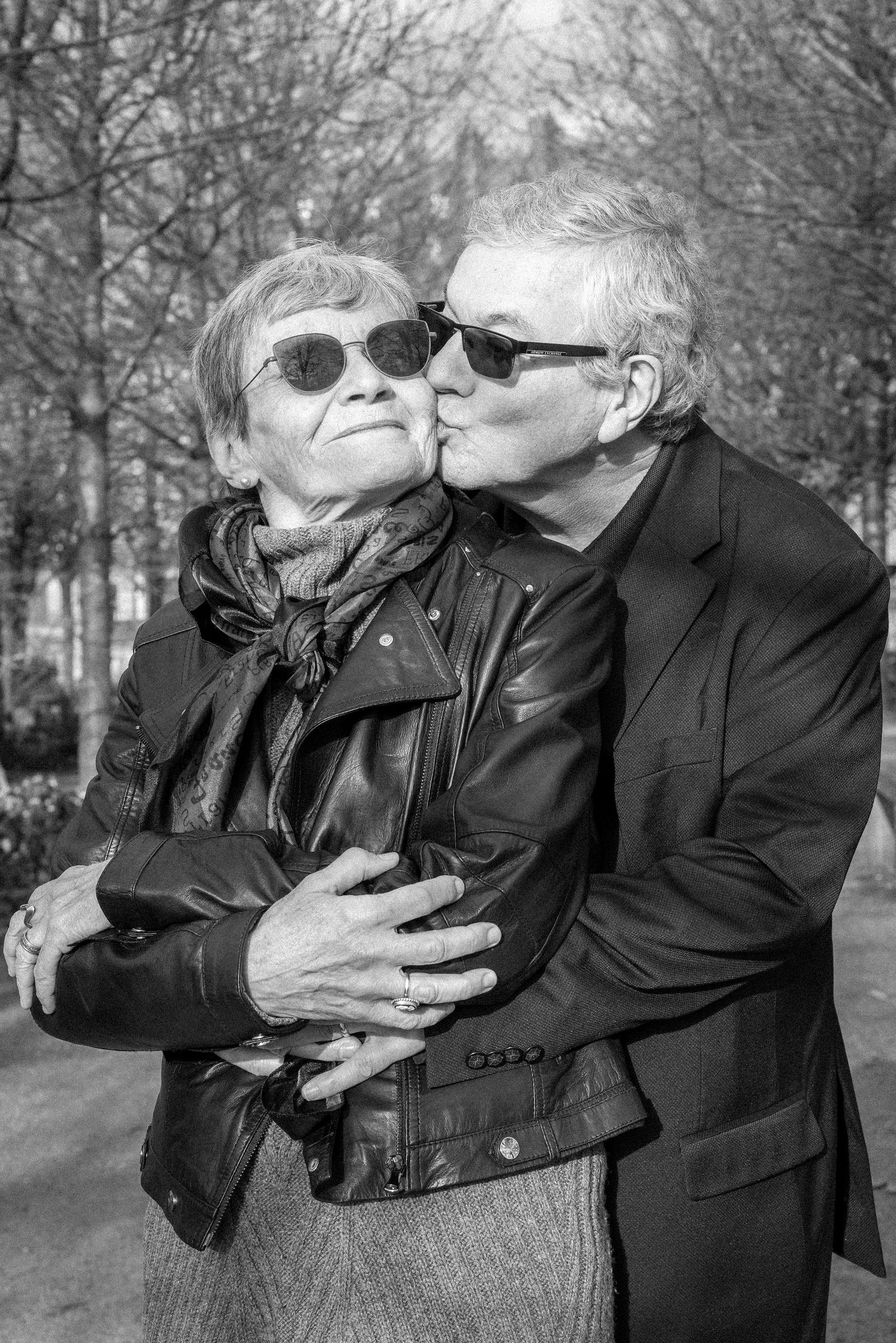 Elderly couple sharing a kiss outdoors, both wearing sunglasses, man embracing woman in black and white photo.