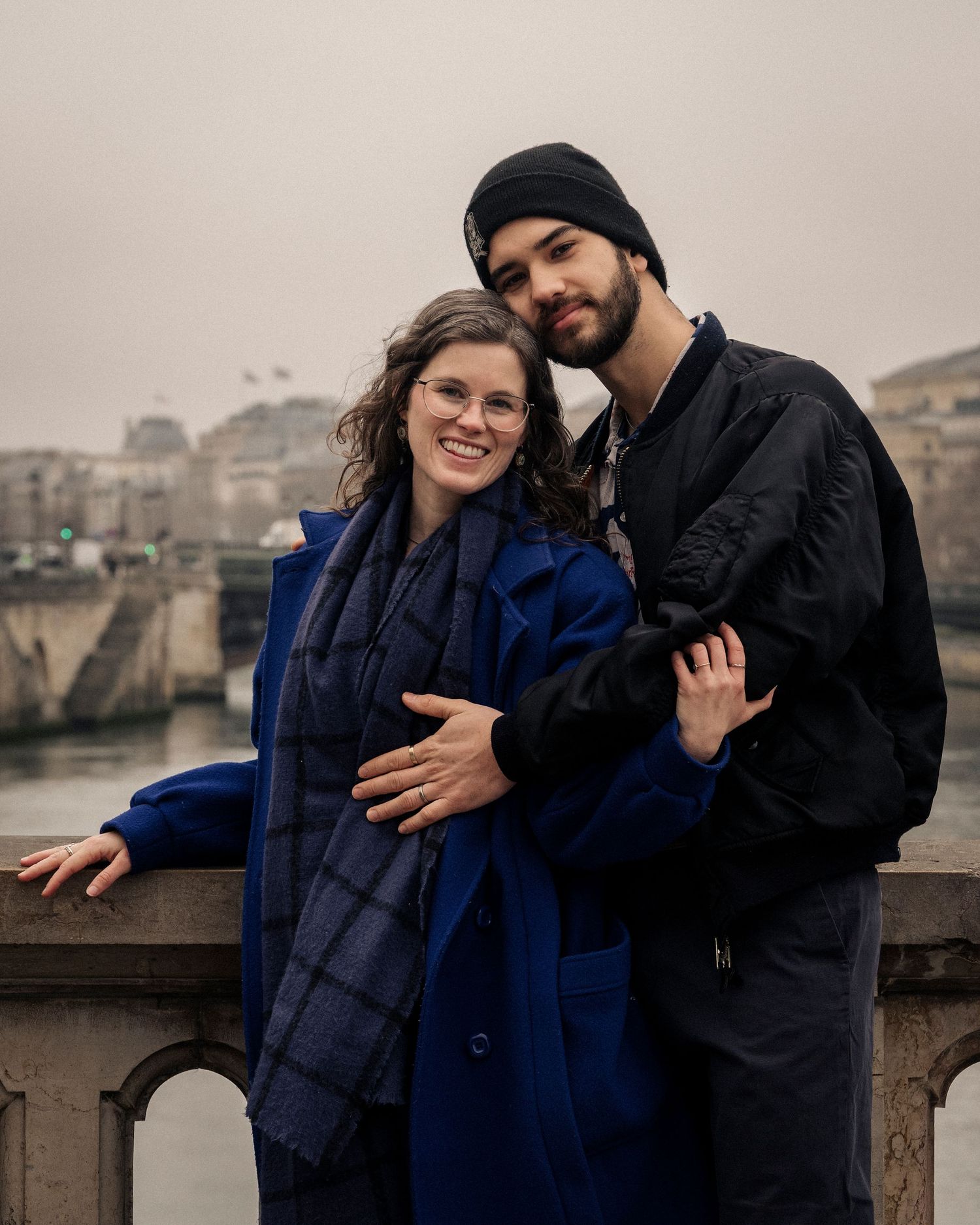 Couple embracing on a bridge in Paris on a cloudy day, woman in blue coat and man in black jacket.