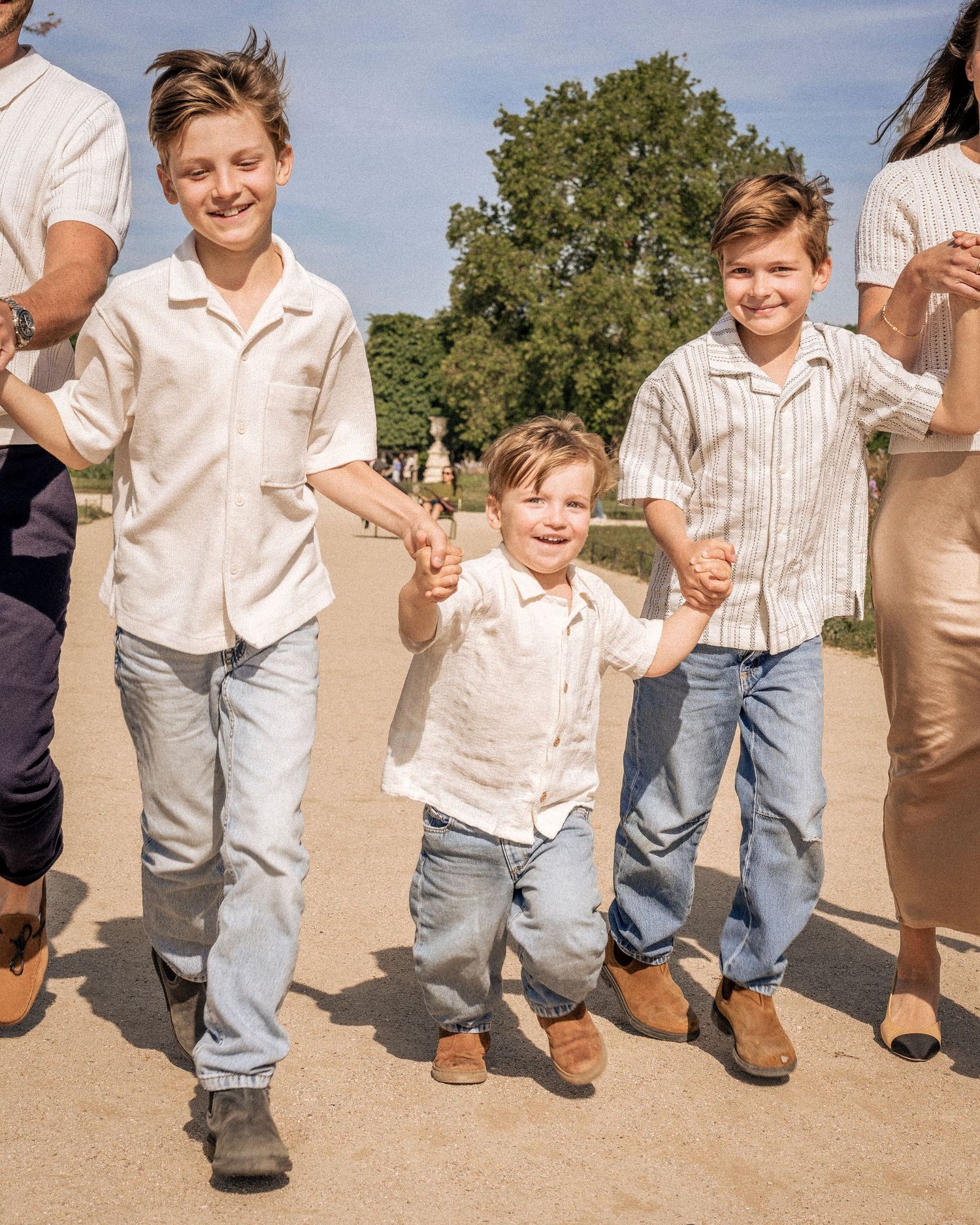 Three young brothers in matching white shirts and jeans holding hands and walking outdoors.