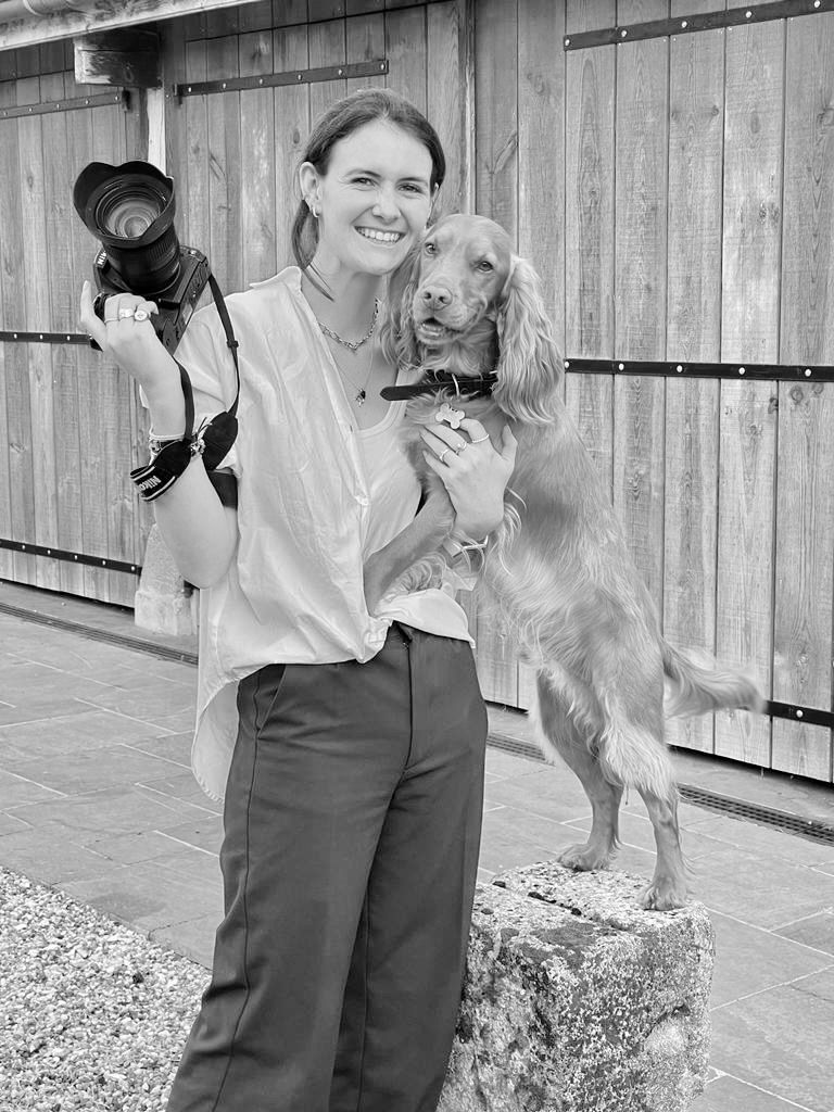 Smiling woman holding DSLR camera and cocker spaniel dog outdoors in black and white portrait.