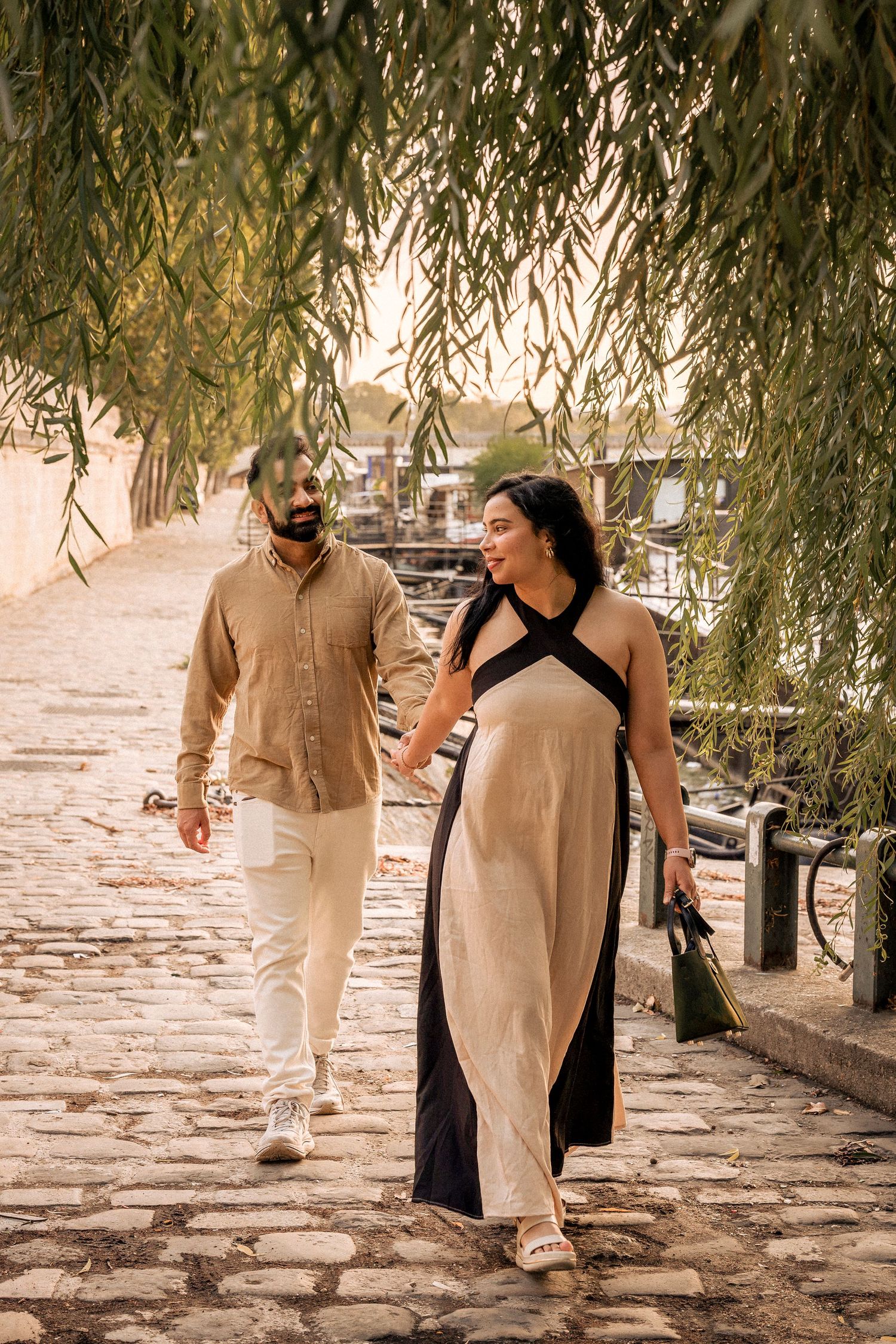 A couple walks hand-in-hand along a cobblestone Paris riverside path beneath weeping willow trees.