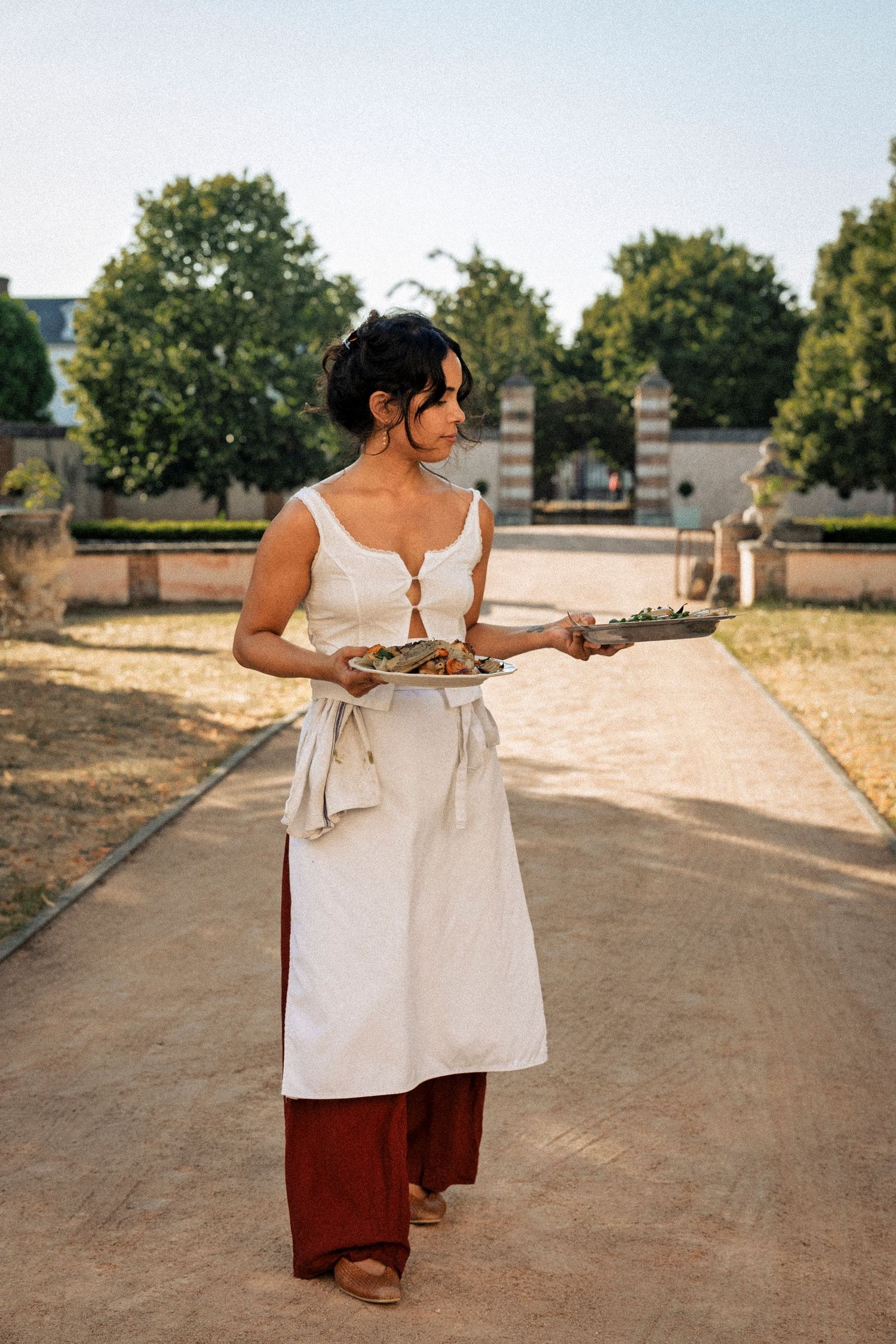 Woman in white apron carrying food platters along a tree-lined garden path in warm sunlight.