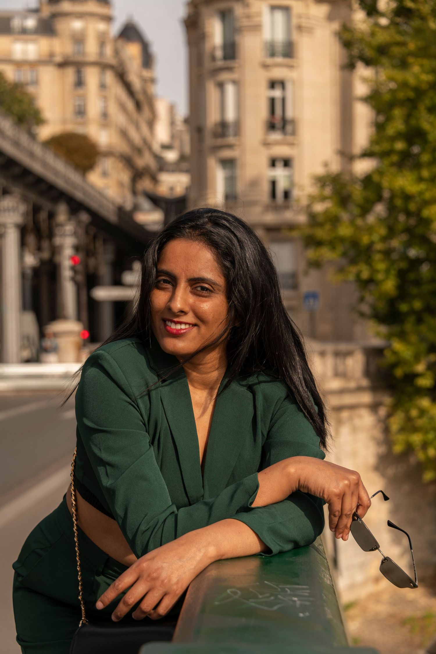 Smiling woman in green blazer leaning on railing with Parisian architecture in background.