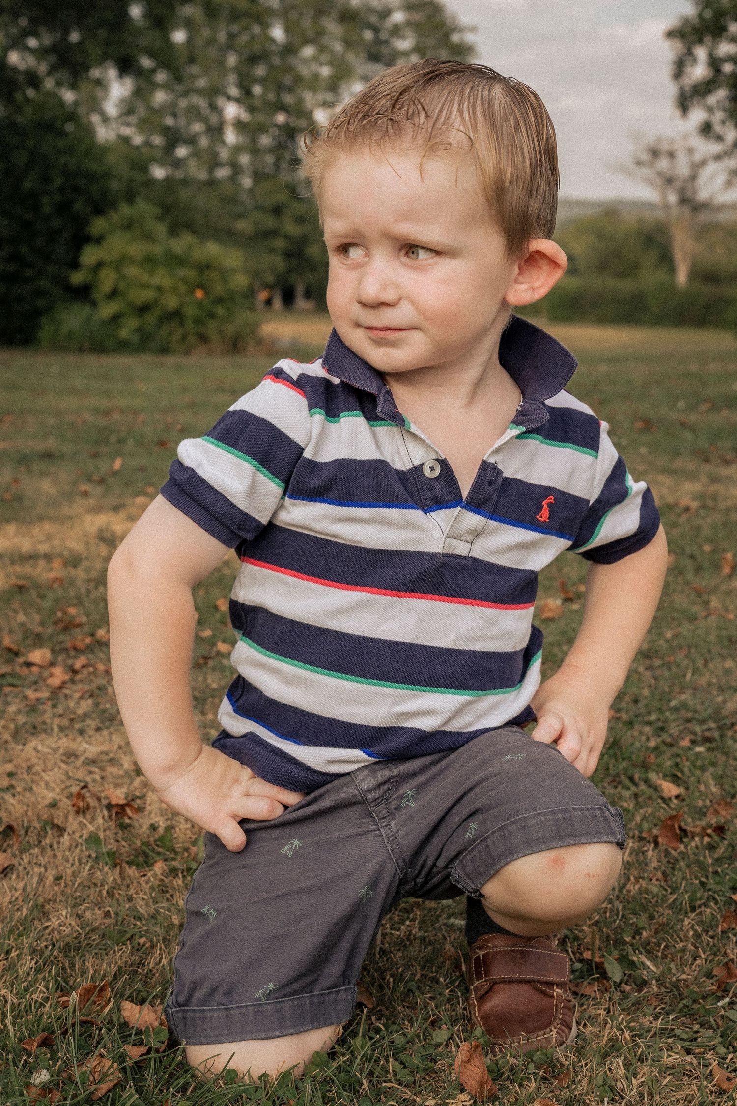 Adorable toddler boy in striped polo shirt sitting on green grass in a park, looking sideways with curiosity.