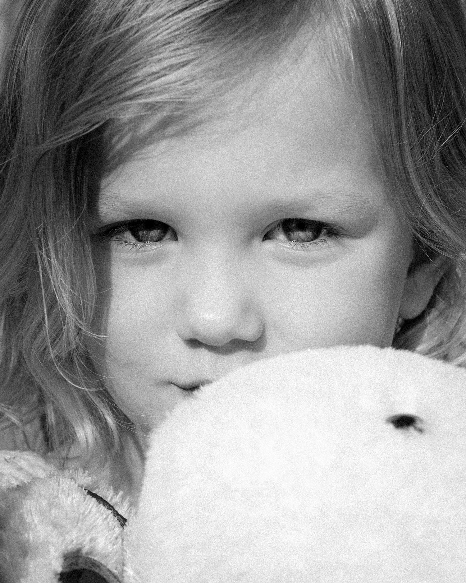 Black and white close-up of a young blonde girl holding a white stuffed teddy bear up to her face.