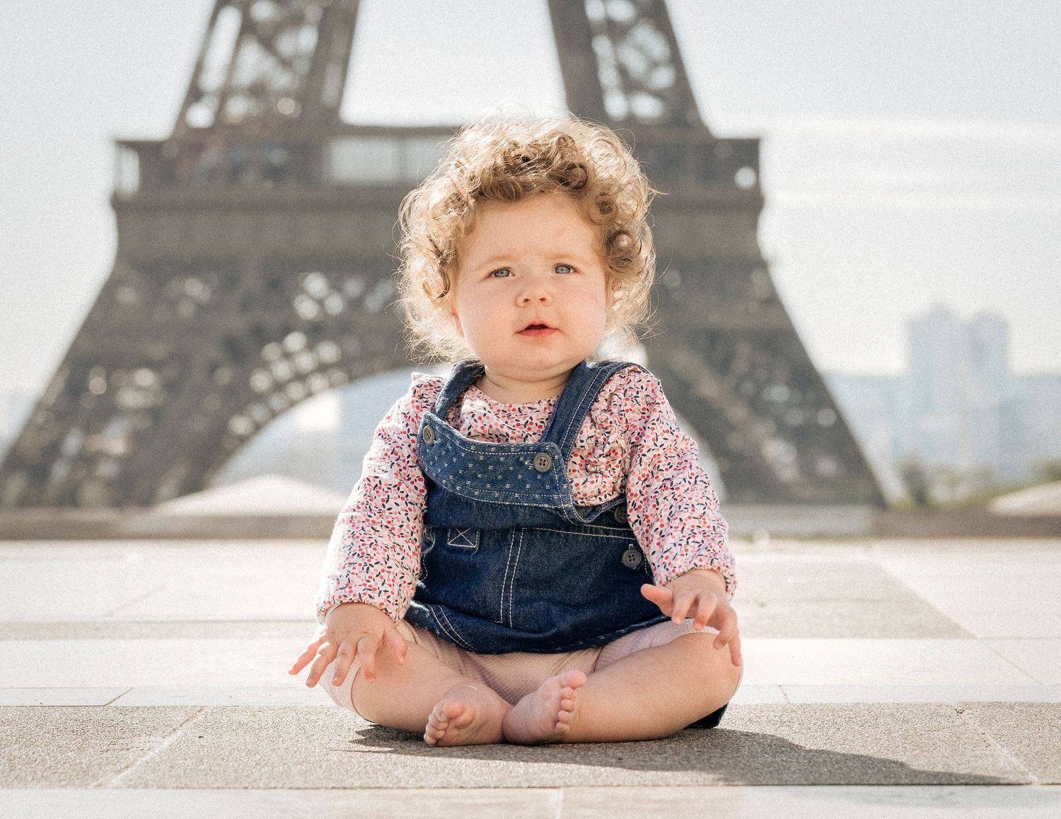 Adorable curly-haired toddler in denim overalls sitting cross-legged with Eiffel Tower behind her.