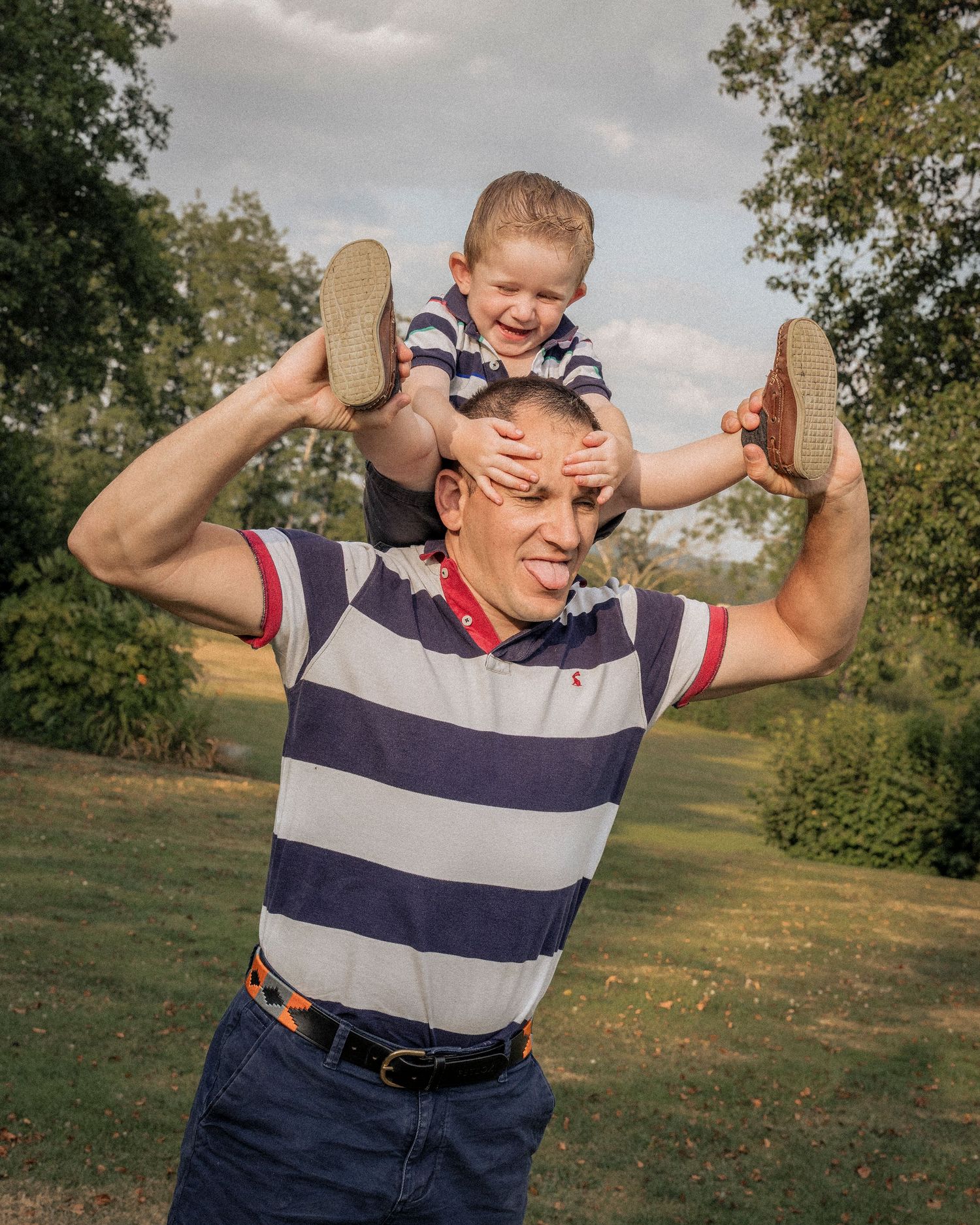 Laughing toddler sits on smiling mans shoulders outdoors in a lush green park on a cloudy day.