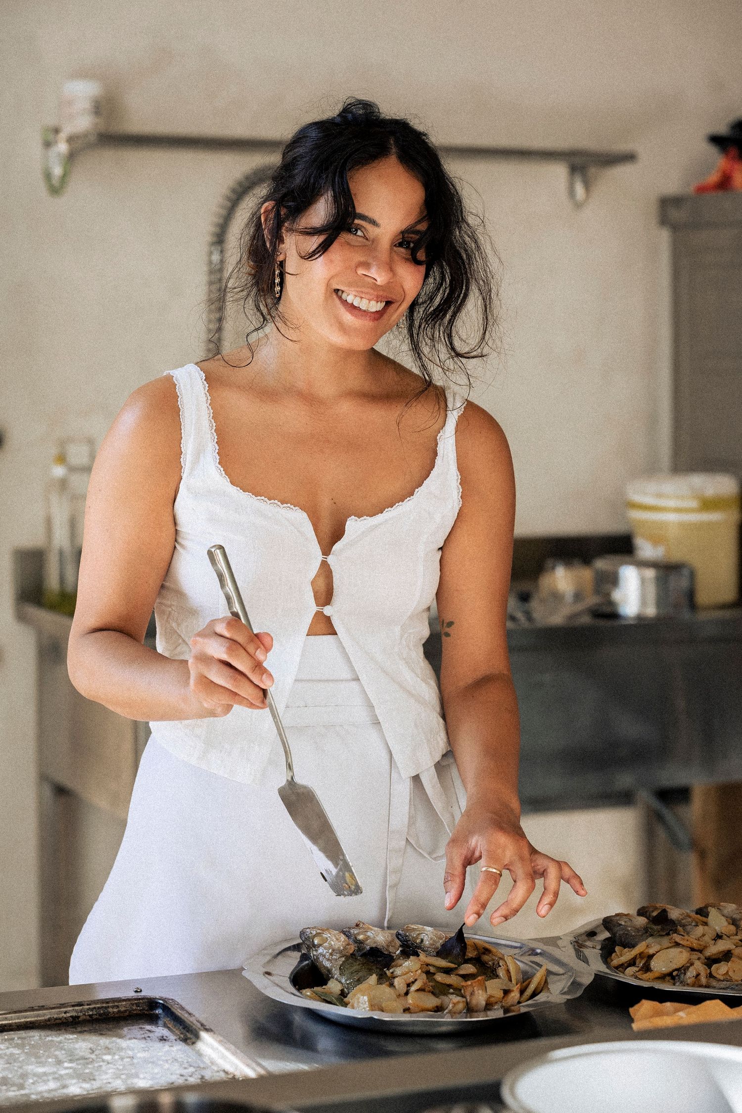 Smiling woman in white top serving food with tongs in a rustic kitchen setting.