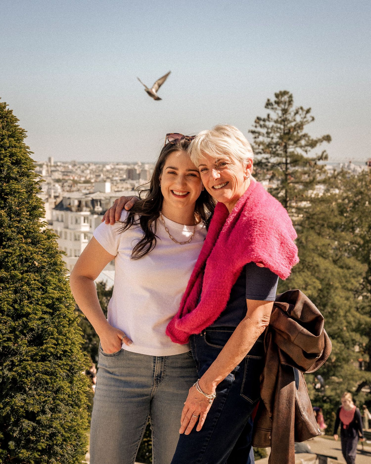 Two women smiling together on a sunny overlook with Paris cityscape in the background.
