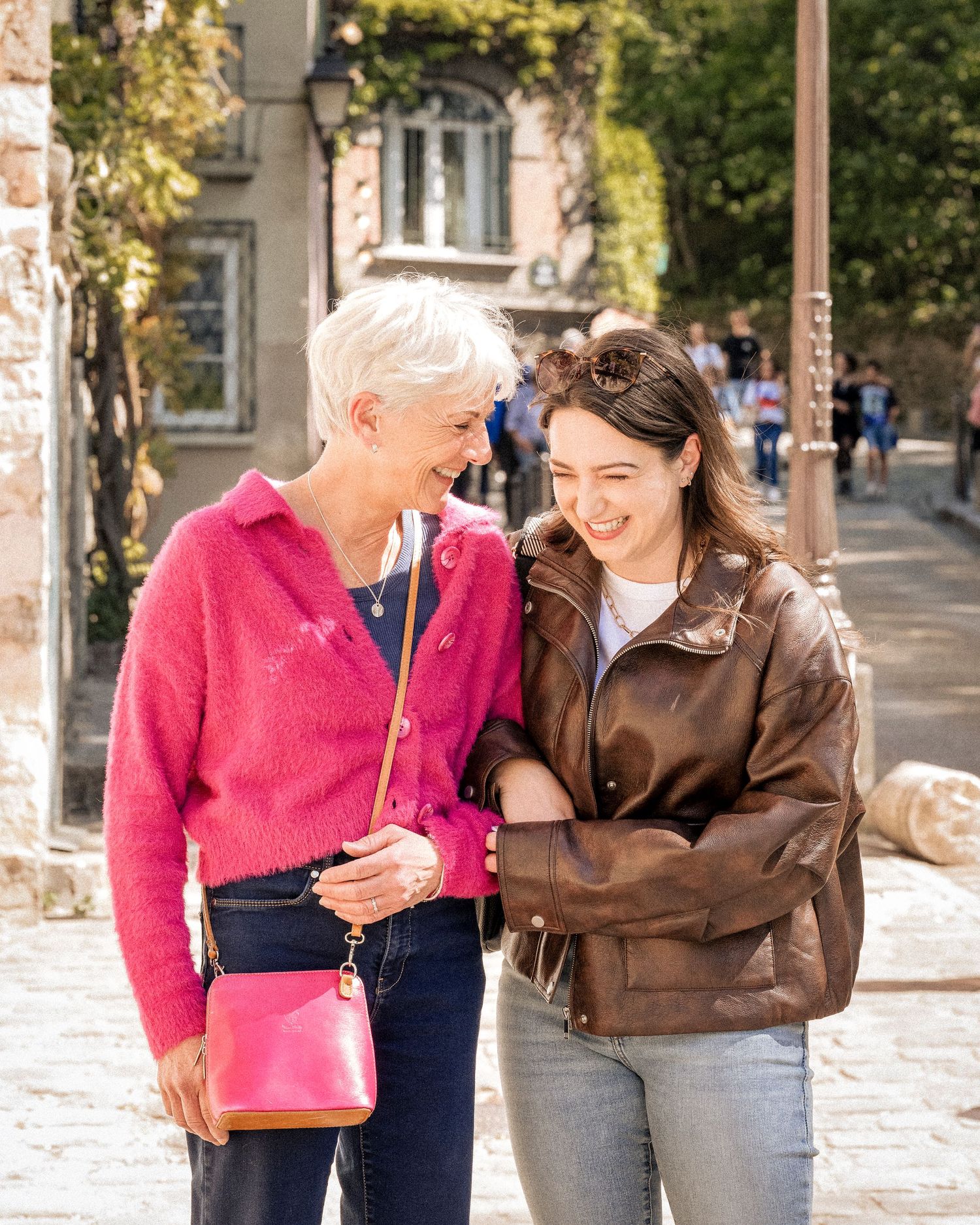 Two women laughing together outdoors, one in a pink sweater with a pink bag, the other in a brown leather jacket.