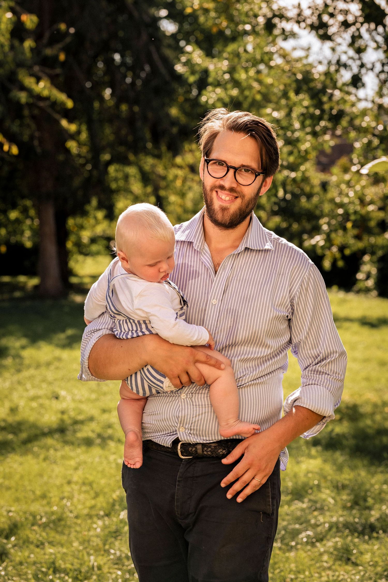 Smiling bearded man wearing glasses holds a baby outdoors in a sunny green park setting.