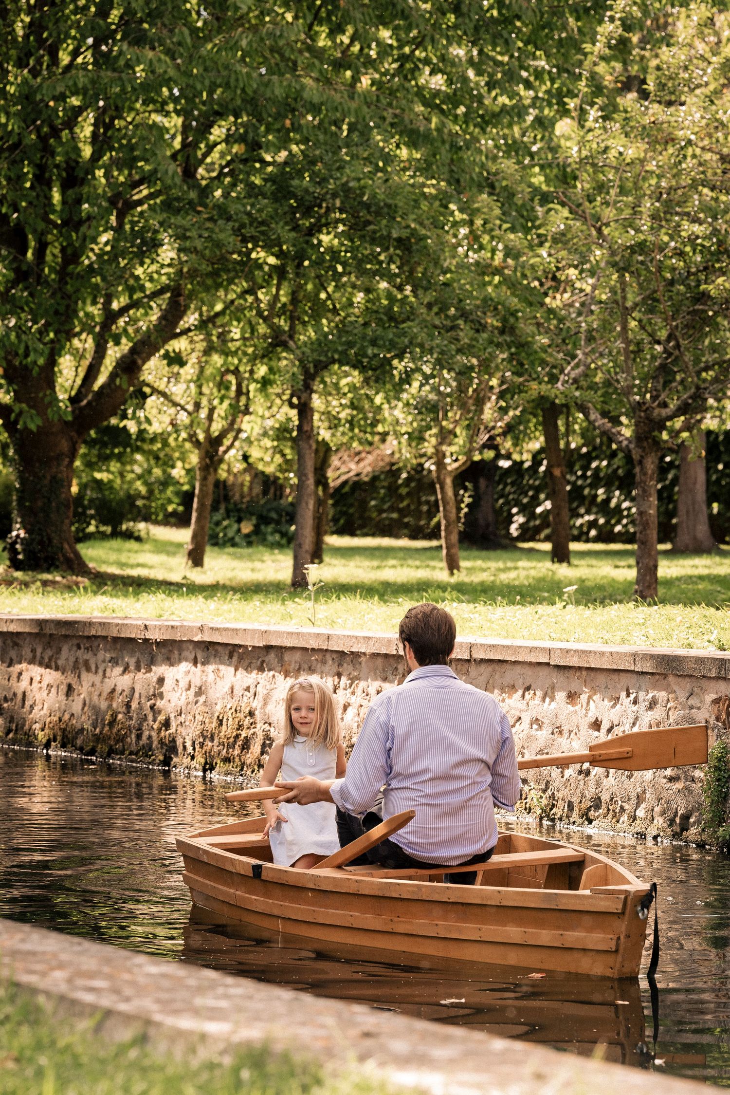 A man and young girl sit together in a wooden rowboat on a calm pond surrounded by lush green trees.