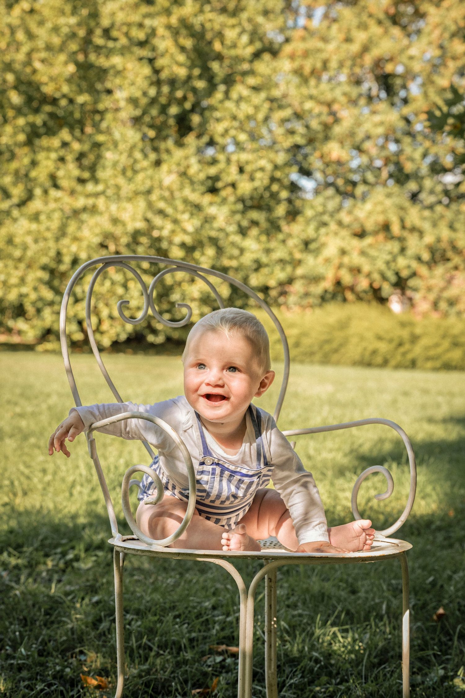 Happy baby sitting in ornate metal chair on green lawn in sunny garden.