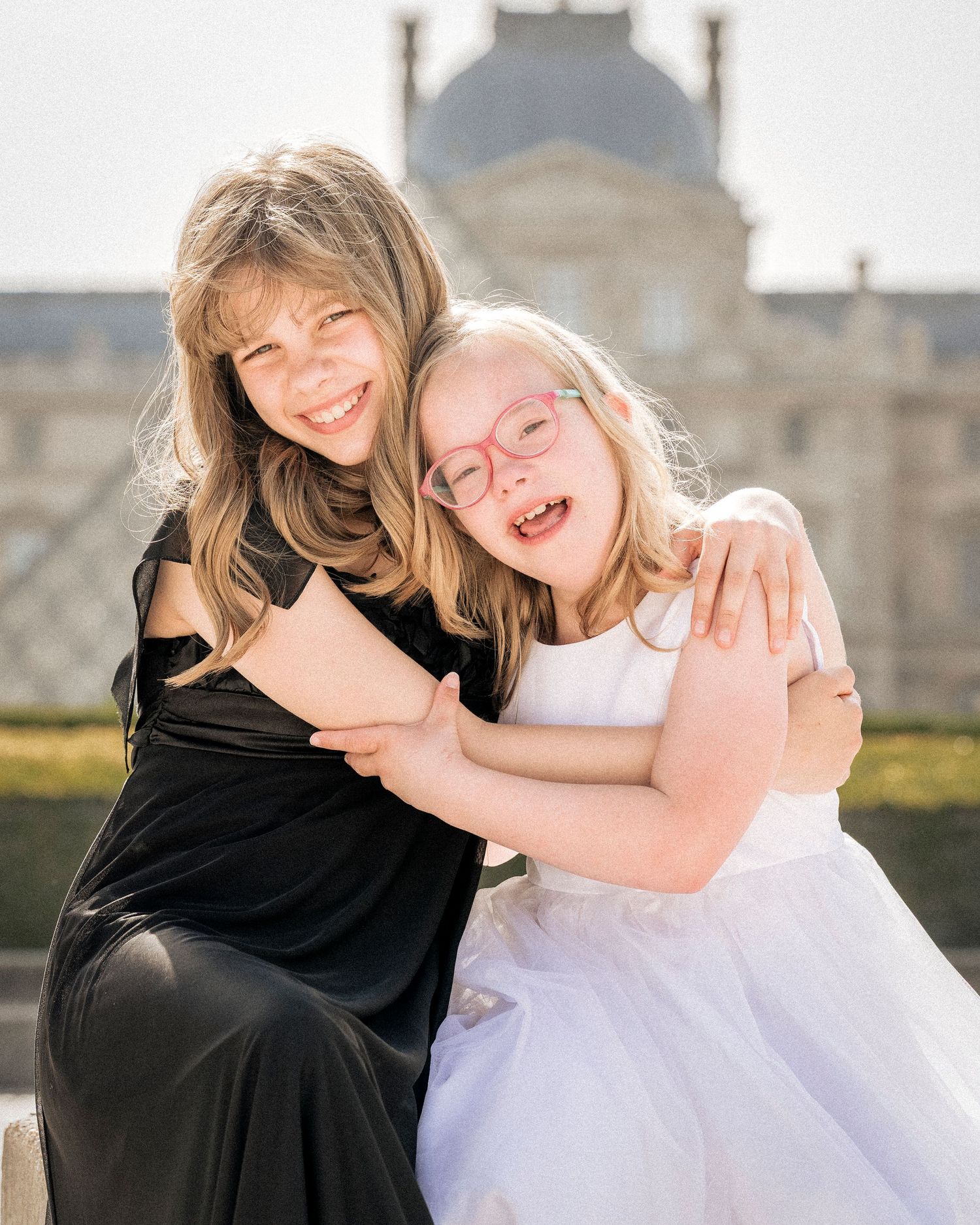 Two smiling girls hugging warmly outdoors near the Louvre in Paris, one in black dress and one in white tulle dress.