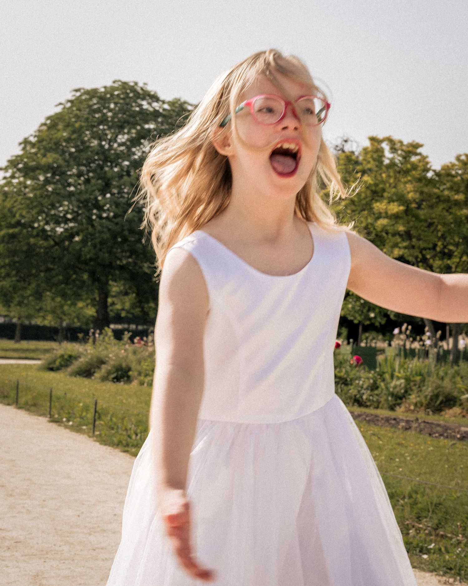 Young blonde girl with pink glasses in white dress spinning joyfully in a sunny park, arms outstretched with excitement.