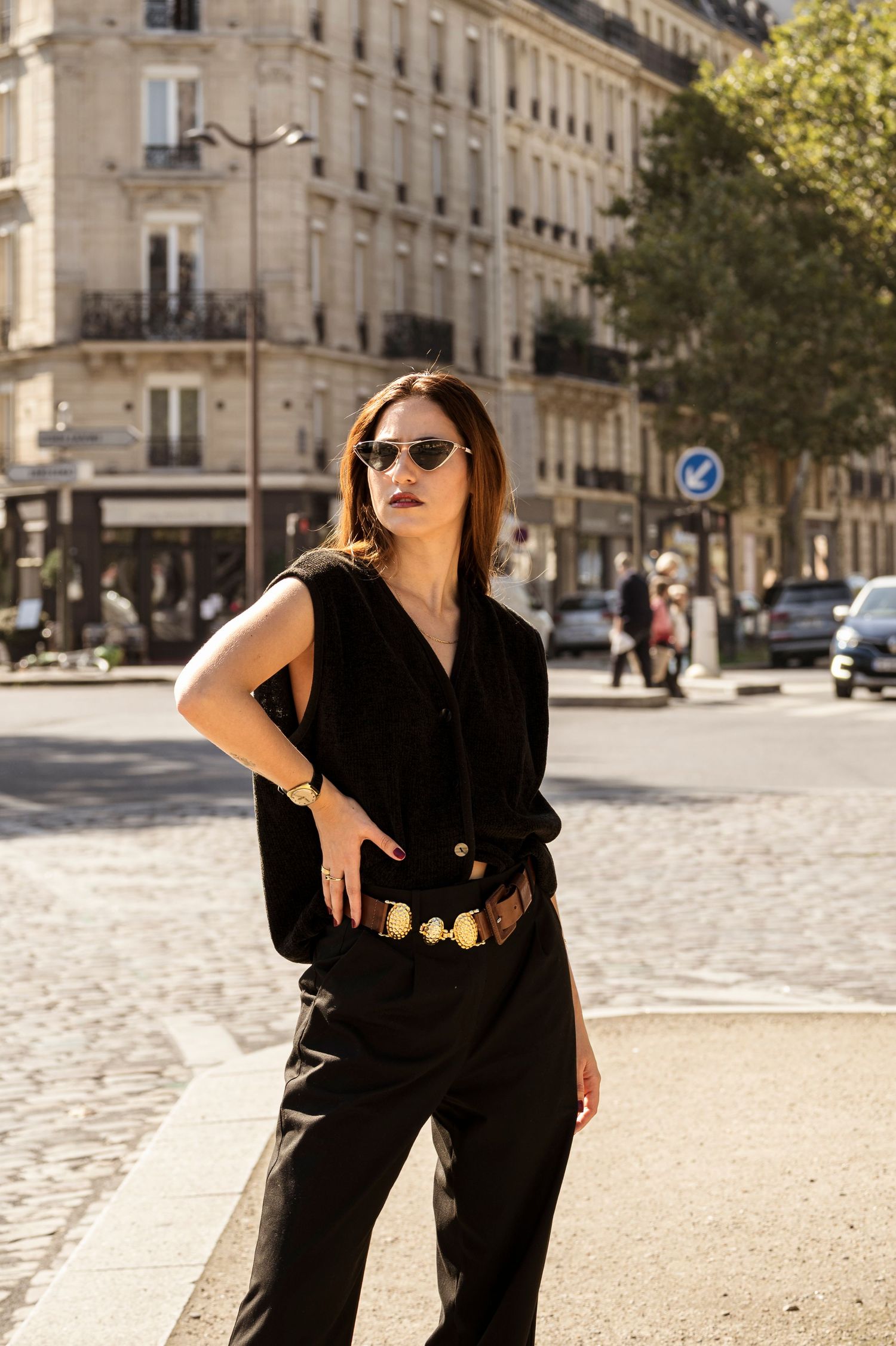 Stylish woman in black outfit with gold belt and sunglasses posing on Parisian cobblestone street.