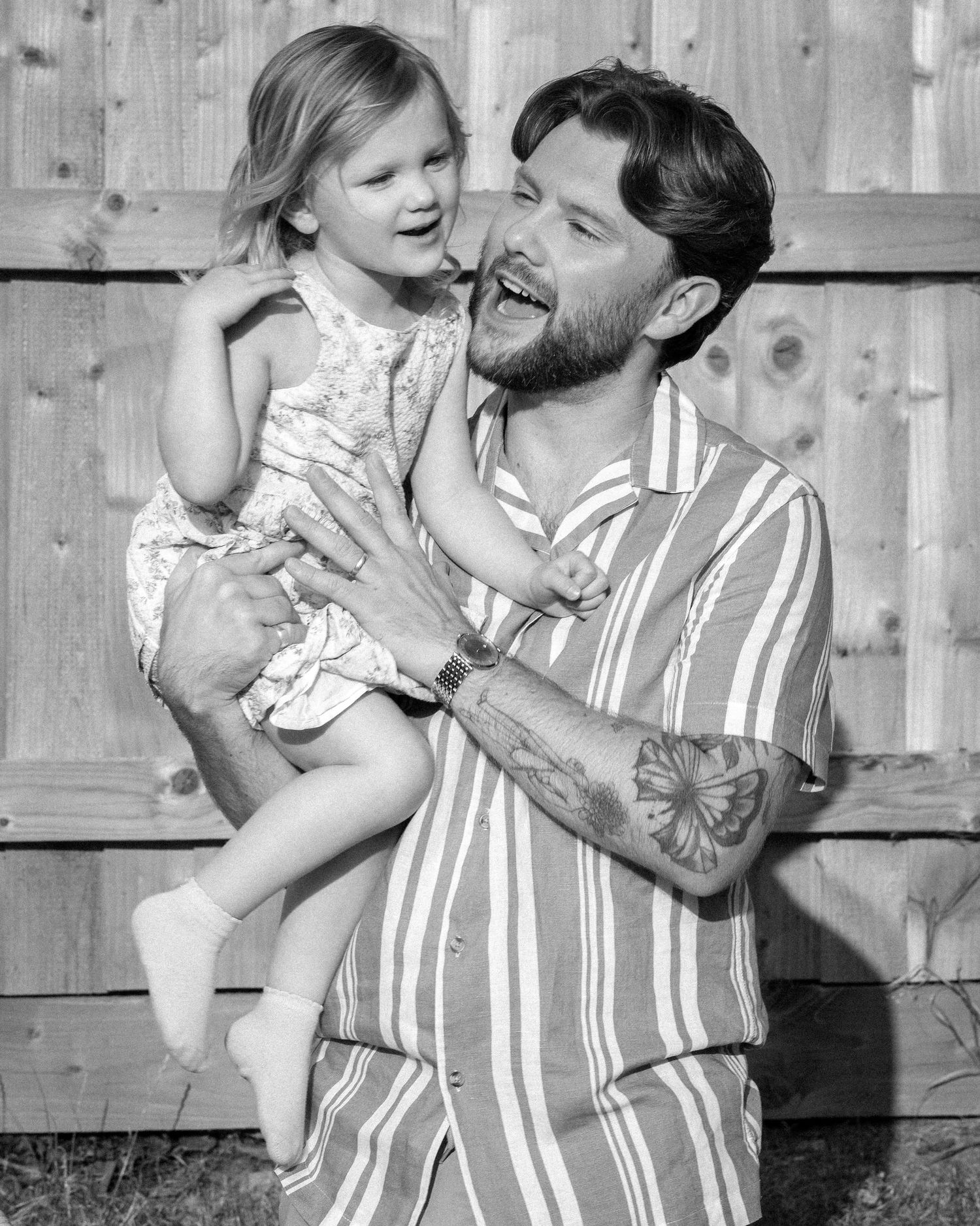 Tattooed man laughing joyfully while holding a smiling toddler girl in black and white photo.