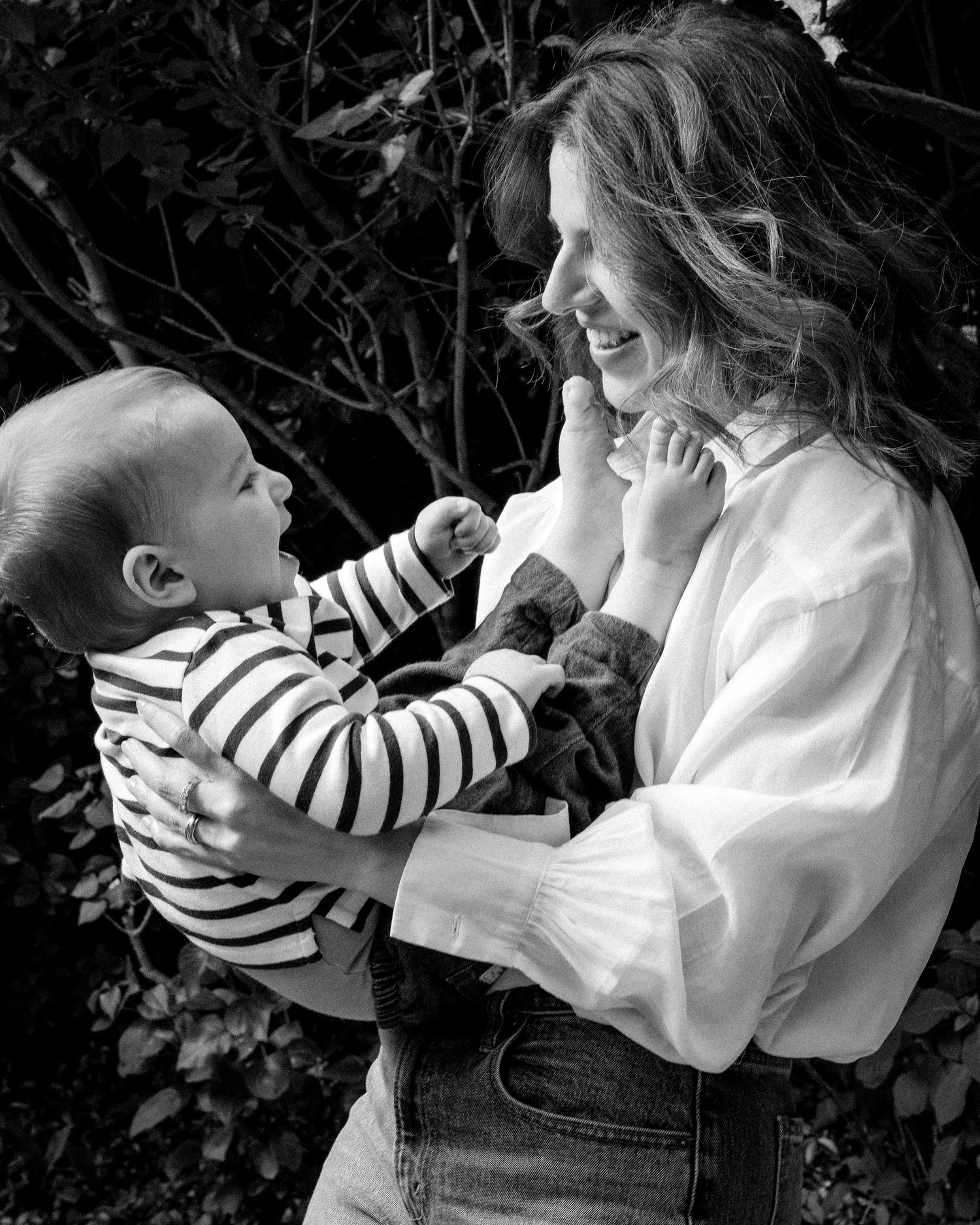 Black and white photo of a smiling mother holding her baby boy in a striped outfit outdoors.