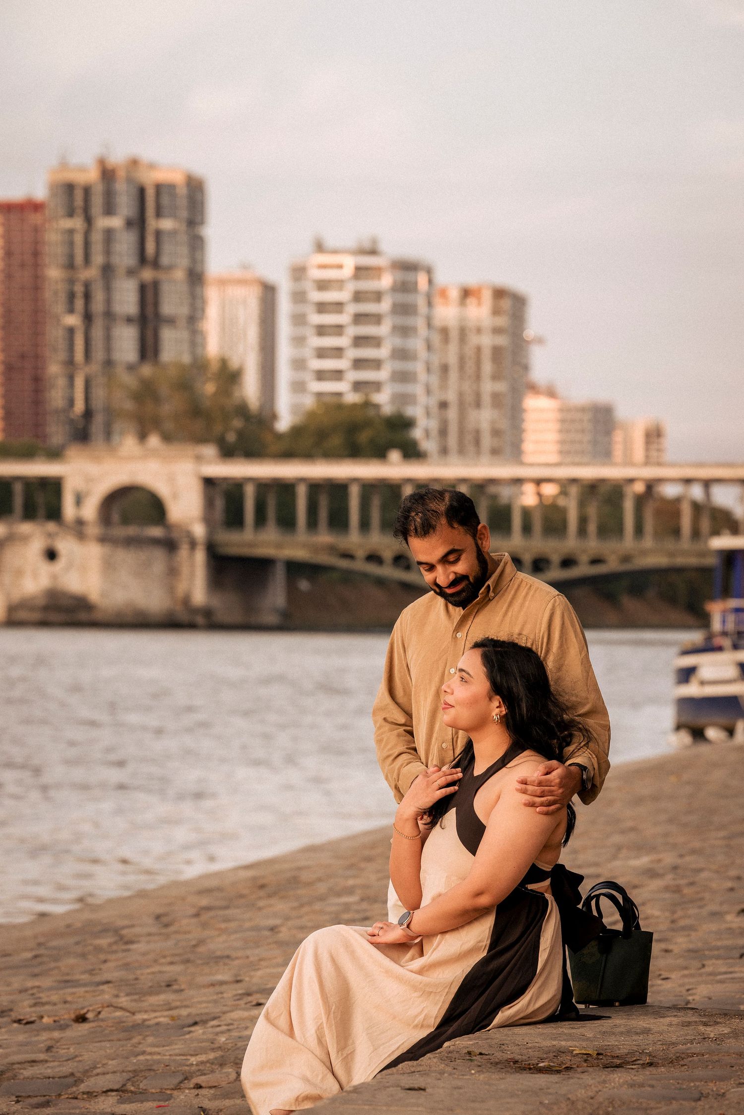 Couple sitting together on riverside cobblestone bank at sunset with city bridge in background.
