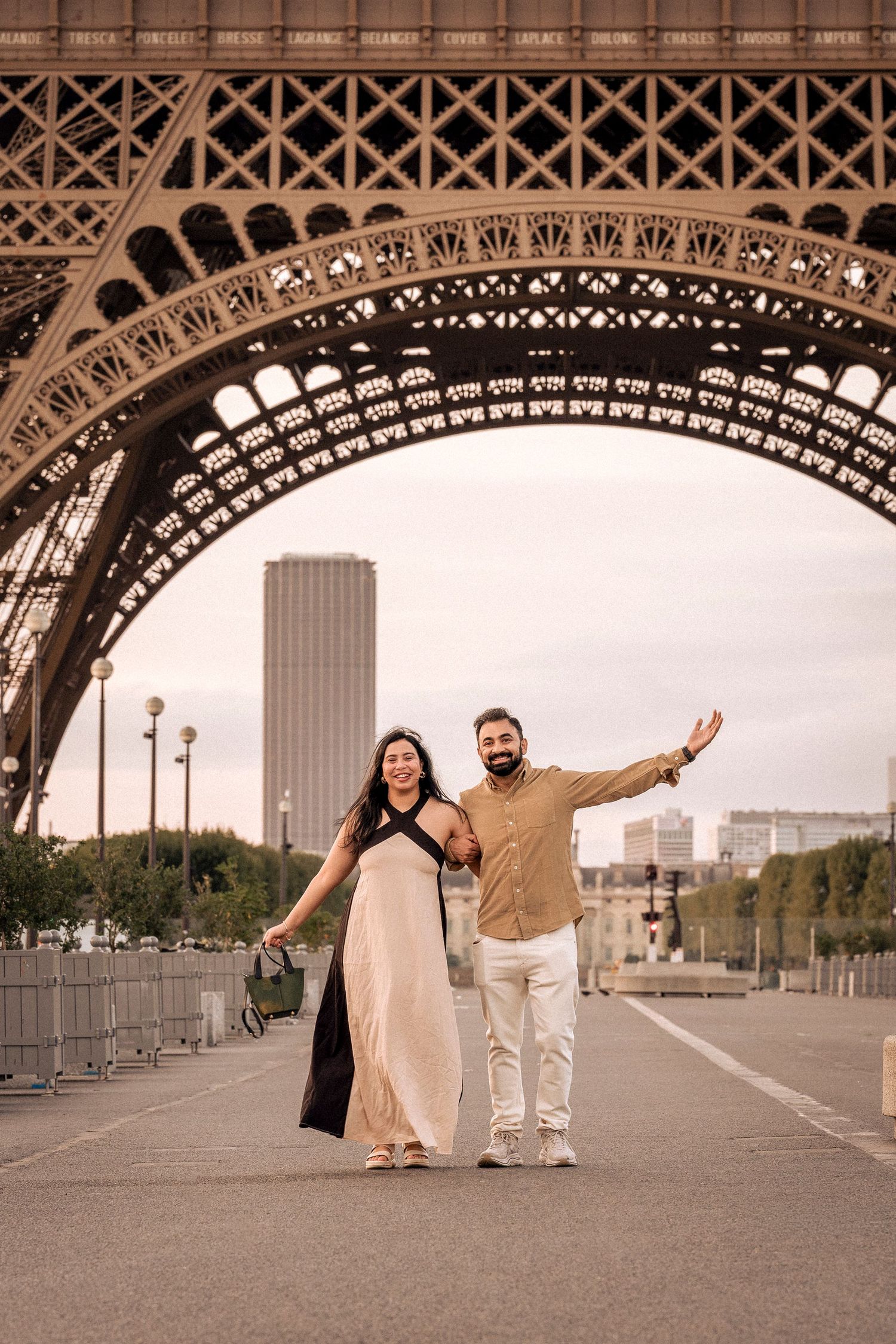 Happy couple walking and waving beneath the iconic Eiffel Tower structure in Paris, France.